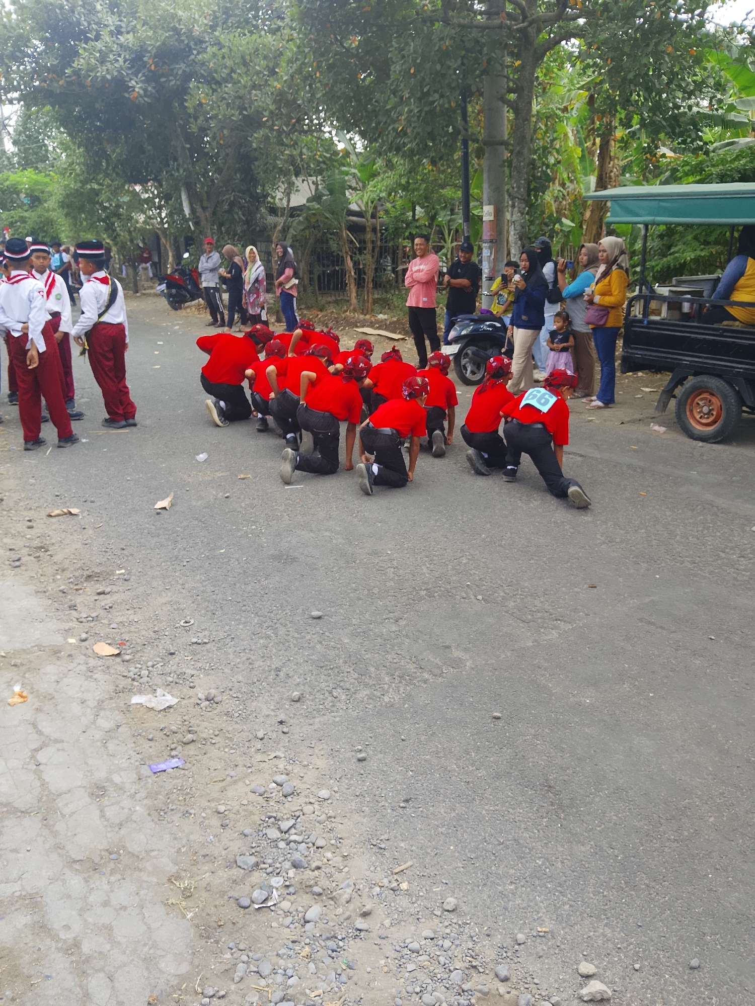 The image depicts a street scene in Bangsri, Indonesia, likely during a parade or event. In the foreground, a group of young individuals wearing red uniforms and red headbands are kneeling on the asphalt road, facing away from the camera. Their uniforms consist of red t-shirts and black pants, with some individuals wearing red helmets or head coverings. One individual in the back has the number "66" visible on their red shirt. To the left, two individuals in white shirts with red pants and black hats, likely part of a different group, are standing. Behind them, more spectators are visible along the side of the road, some standing and others seated. A black truck with a green canopy is parked on the right side of the road, with people standing around it. The environment includes a paved road with scattered debris, trees lining the sides of the road, and buildings in the background. The overall atmosphere appears to be that of an outdoor community gathering.