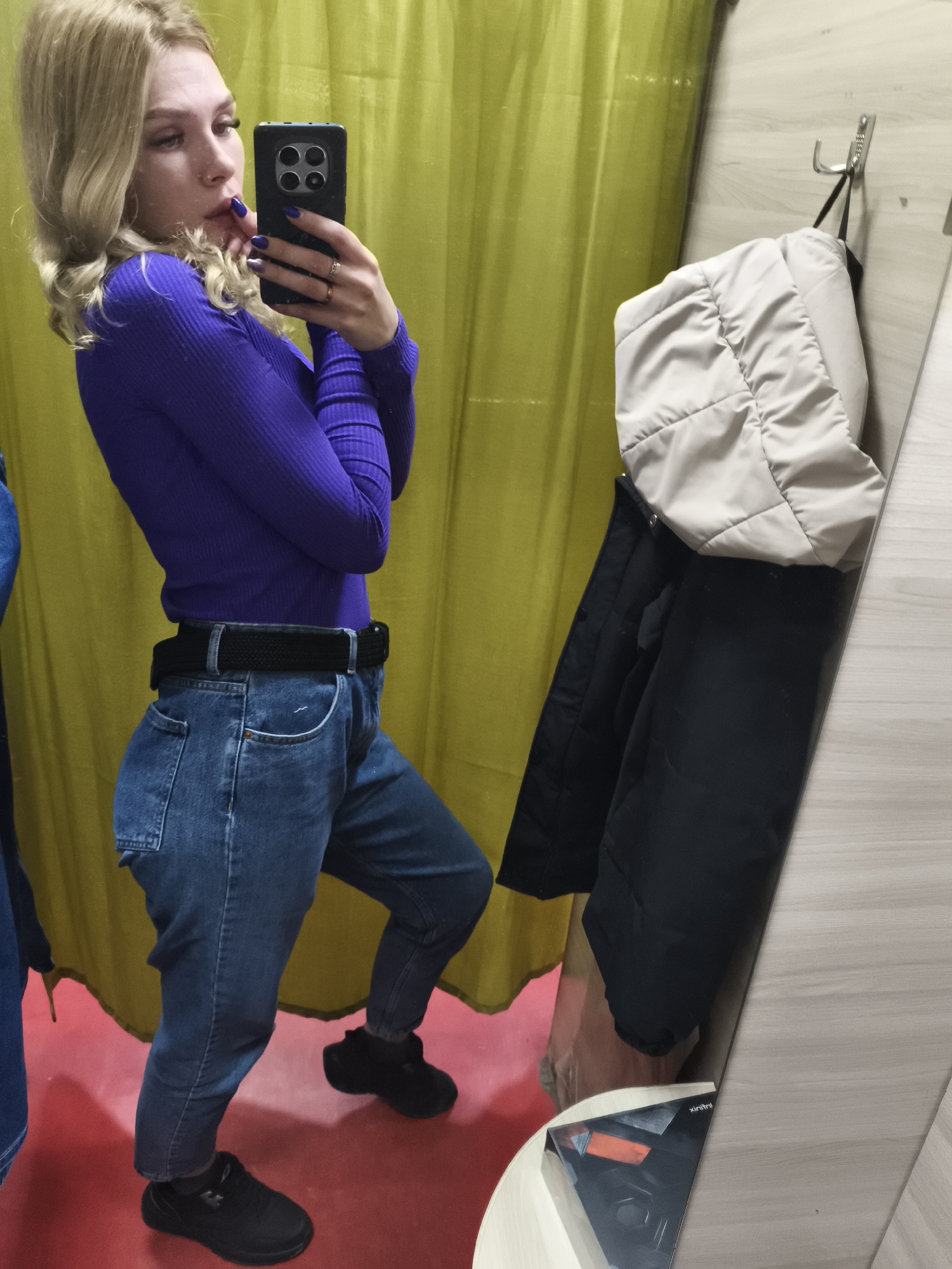 A young blonde woman is taking a selfie in a fitting room. She is wearing a purple long-sleeved shirt, blue jeans with a black belt, and black shoes. She is holding a black smartphone. The fitting room has a yellow curtain as a backdrop, a red floor, and a mirror reflecting the scene. A coat is hanging on a hook next to the mirror. A sticker with text is placed on the bottom left corner of the mirror. The fitting room is potentially located in a store in Izhevsk, Russia.