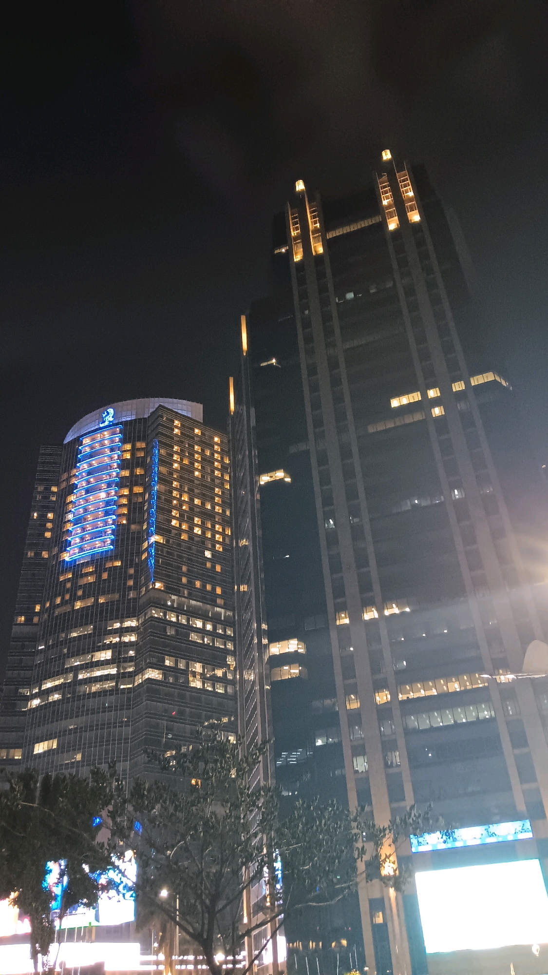 This is a nighttime photograph taken in Central Jakarta, Indonesia, showcasing two tall skyscrapers illuminated by artificial lights against a dark sky. The skyscraper on the left has a curved top and features a prominent blue light display on its facade, spelling out a logo. Numerous windows across both buildings are lit with warm yellow light. The skyscraper on the right is taller and more angular, with vertical strips of warm yellow light accentuating its structure. In the foreground, silhouetted trees partially obscure the lower sections of the buildings, and the glow from street-level lights and digital billboards is visible at the very bottom of the frame. The overall scene depicts an urban nighttime environment with architectural illumination as the primary focus.