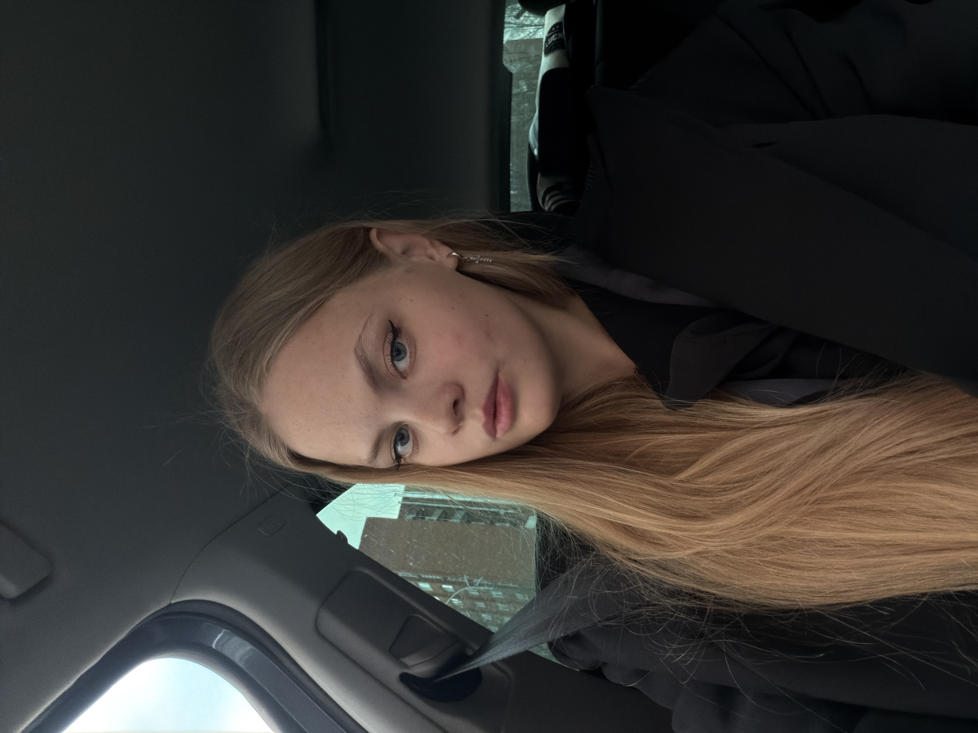 A young woman with long, straight blonde hair, light blue eyes, and fair skin is pictured inside a car. She is resting her head against a surface, possibly the car's window frame, and gazing directly at the camera with a calm, neutral expression. She is wearing a black jacket or top, and a small silver-colored cross earring is visible in her right ear.
The setting is the interior of a vehicle, likely the passenger side, with parts of the car's headliner, window frames, and a deployed seatbelt prominently visible across the lower-left portion of the frame. Through the car window, an urban environment can be seen in the background, including a building with multiple stories, suggestive of residential or commercial architecture. Faint white specks on the window or in the background might indicate light precipitation such as snow or rain.
The time of day appears to be daytime, though the light is diffused, suggesting an overcast sky. Notable details include an "AIRBAG" label visible on the car's interior above her head, and partially visible text that spells "LUCK" on an object or piece of clothing seen through the window in the background. The location context for this scene is Izhevsk, Russia.