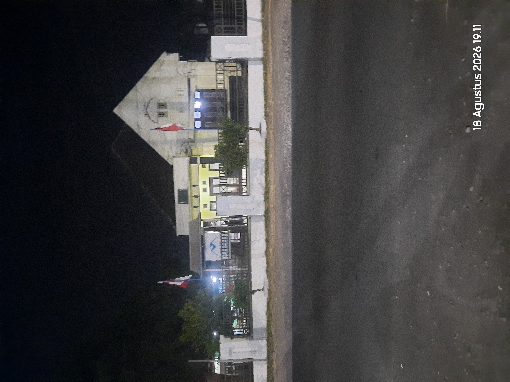 The image depicts a building at night, likely a residence or public building, located in Sukokerto, Indonesia. A red and white flag is prominently displayed on a flagpole in front of the building. The building itself is light-colored with dark windows and appears to have two stories. A fence with white posts runs along the front of the property, separating it from a road in the foreground. The scene is illuminated by artificial lighting, casting shadows and highlighting the architectural features of the building. The sky is dark, indicating it is nighttime. A date stamp in the bottom right corner reads "18 Agustus 2026 19.11".