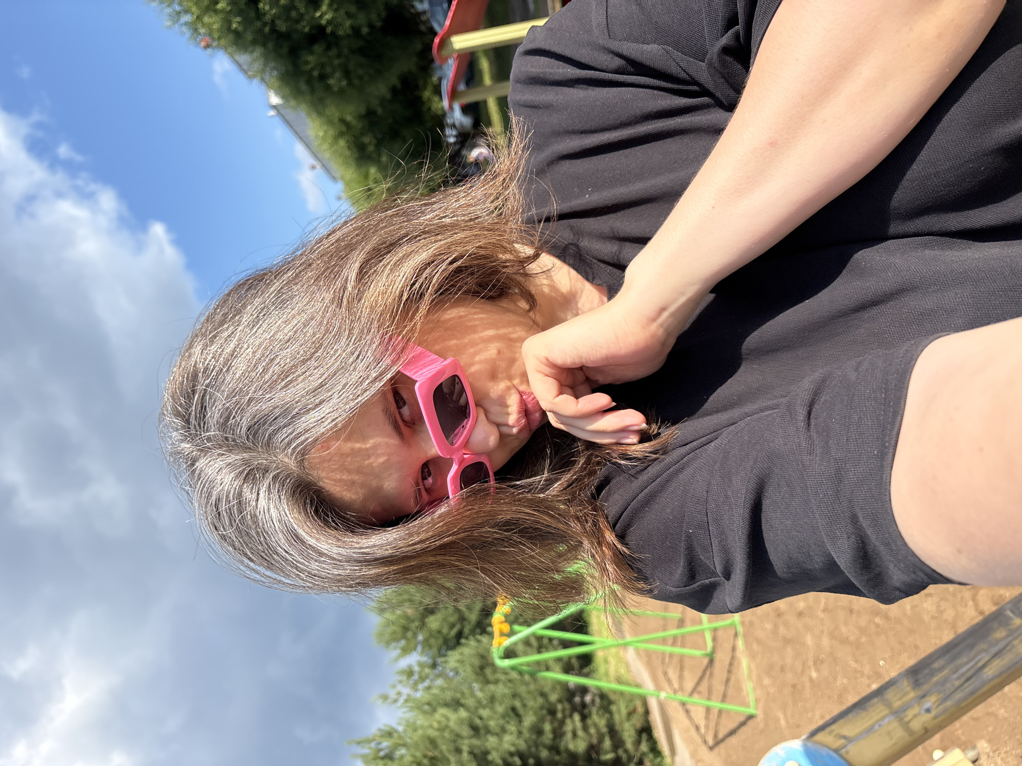 A young woman with long, brown and graying hair, wearing bright pink rectangular sunglasses and a black t-shirt, is taking a selfie outdoors on a sunny day. She is puckering her lips and touching her chin with her right hand. The background shows a playground with a green swing set and brown sand. In the distance, there are green trees and a building. The scene appears to be in a residential area of Chaikovsky, Russia.