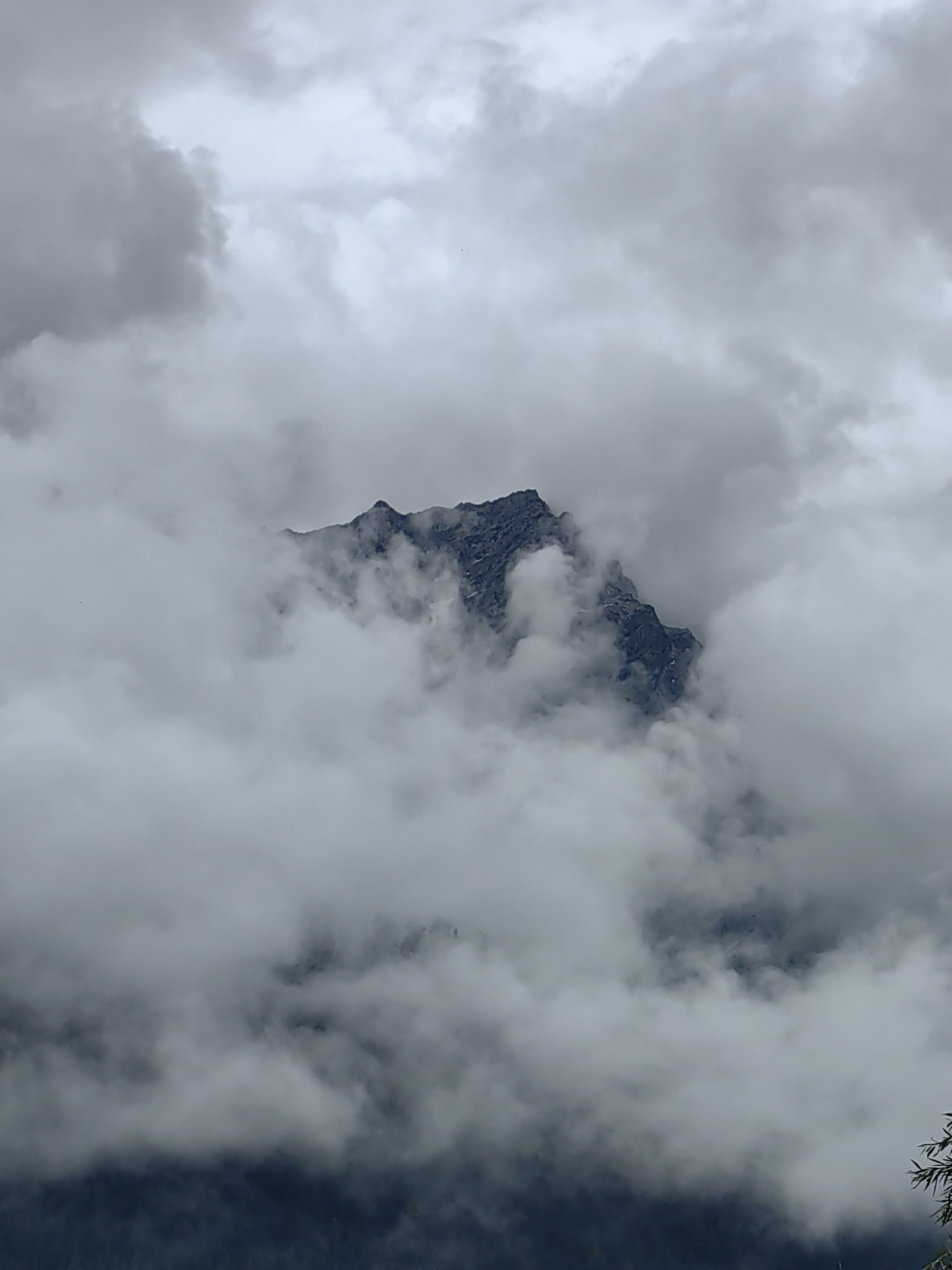 A jagged, dark mountain peak pierces through a thick layer of grey clouds, partially obscuring its rugged slopes. The clouds are dense and varied in texture, with some areas appearing wispy and translucent, while others are solid and opaque. The overall atmosphere is moody and dramatic, suggesting a cool, possibly wet, environment. The mountain itself appears to be made of dark rock, with visible fissures and sharp edges. The image does not contain any people, objects, or actions. The location in Vashisht, India is inferred from the surrounding context provided. The scene is likely a natural landscape.