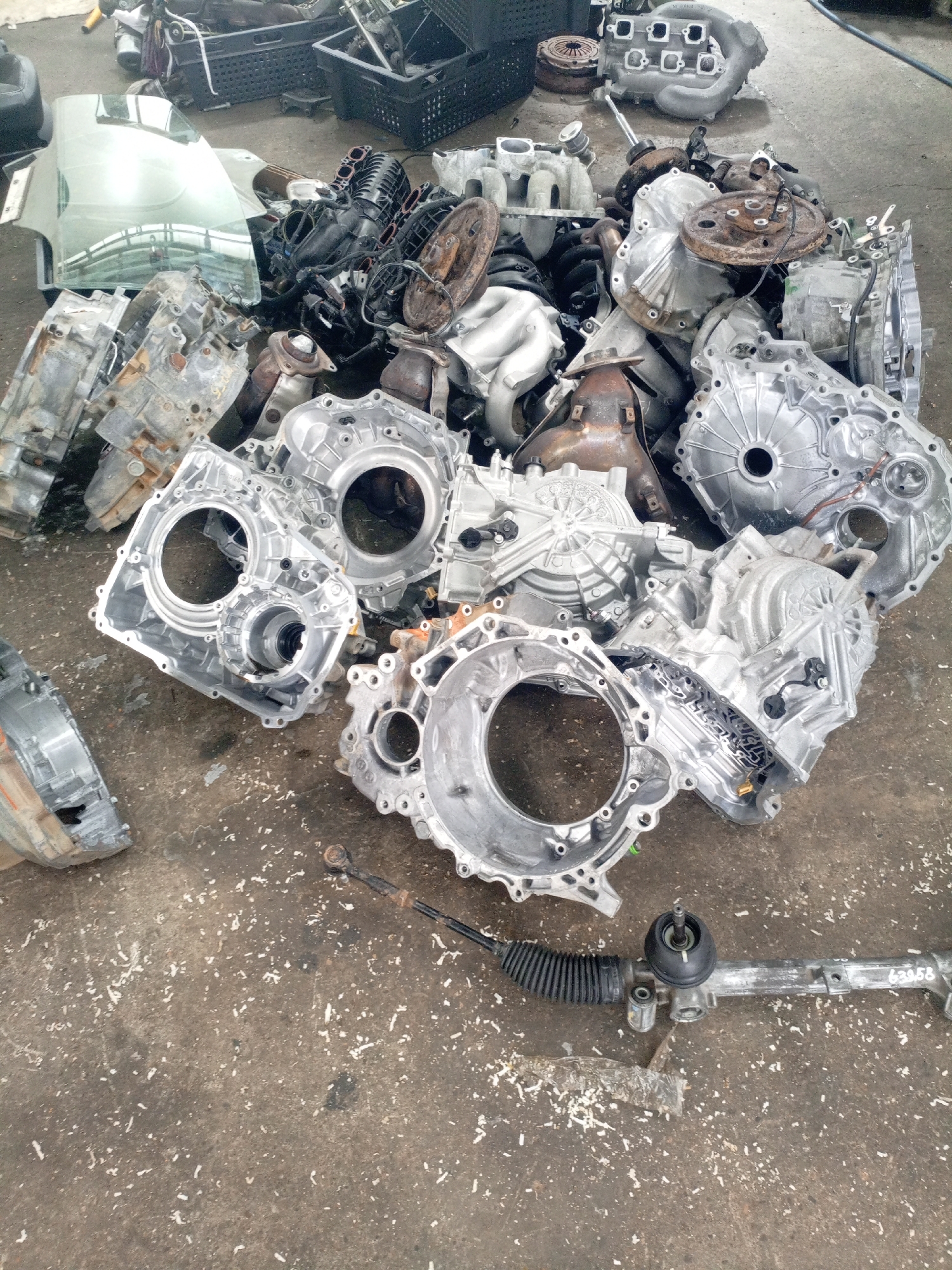 The image depicts a junkyard or auto salvage yard in Oktyabr'skii, Russia. The ground is concrete and littered with various automotive parts, primarily transmissions and engine components, which are scattered in a haphazard pile. Some of these parts are made of aluminum and appear to be somewhat aged and dirty. In the background, there are black plastic crates and other scrap metal. In the foreground, a steering rack with a protective bellows is visible. There are no people or animals present in the image. The overall impression is one of disarray and the accumulation of discarded vehicle parts.
