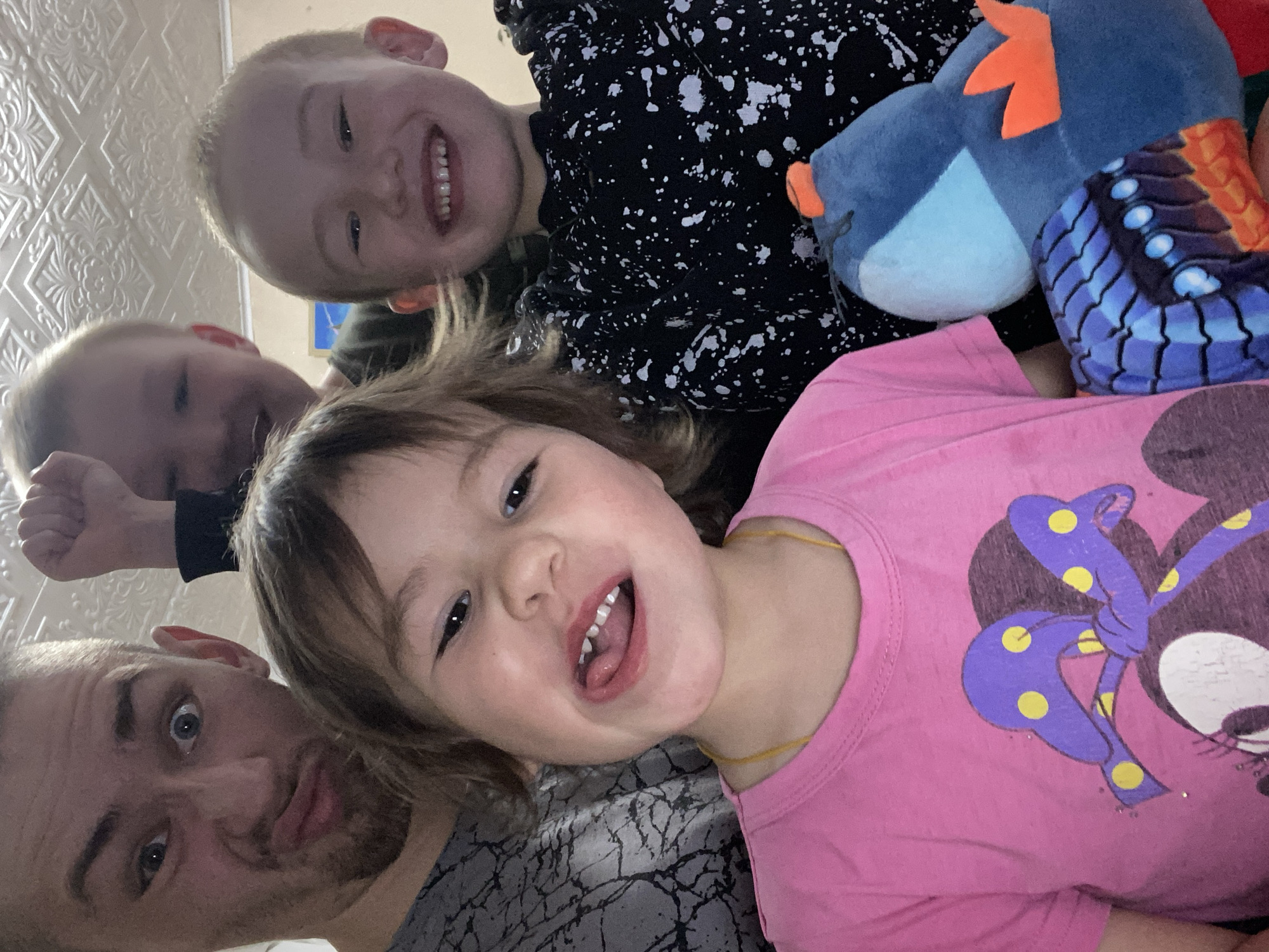 A selfie photograph, possibly taken in a residential setting (given the wall texture visible), features a man and three children. The man is positioned in the lower-left corner of the frame, displaying a surprised expression. One child is in a pink Minnie Mouse shirt. Another child is wearing a black and white splatter-patterned shirt. A blue stuffed animal is also visible. The image provides insufficient detail to confirm a location in the city of Bely Klyuch, Russia. The photo appears to capture a casual, familial moment.
