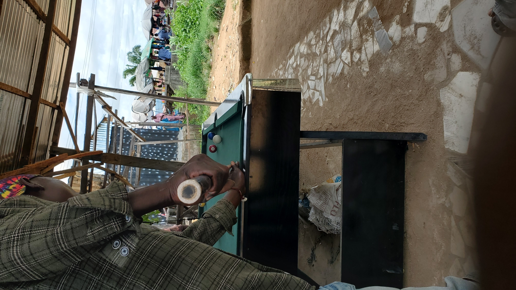 An outdoor billiards table is set up under a corrugated metal roofed shelter. The table is dark green with a black base and is supported by metal legs. Three billiard balls are visible on the table: a red and white striped ball, a solid white ball, and a solid blue ball. A person, wearing a green plaid shirt and a colorful headscarf, is holding a billiard cue over the table, poised to strike a ball. Their hands are positioned on the cue, suggesting they are playing the game. In the background, beyond the shelter, there is a bustling outdoor market scene. Several people are visible, some near stalls covered by umbrellas, and lush green vegetation lines the area. The ground around the table is a mixture of sand and irregular flagstone paving. The overall impression is of a casual recreational area within a vibrant community marketplace.