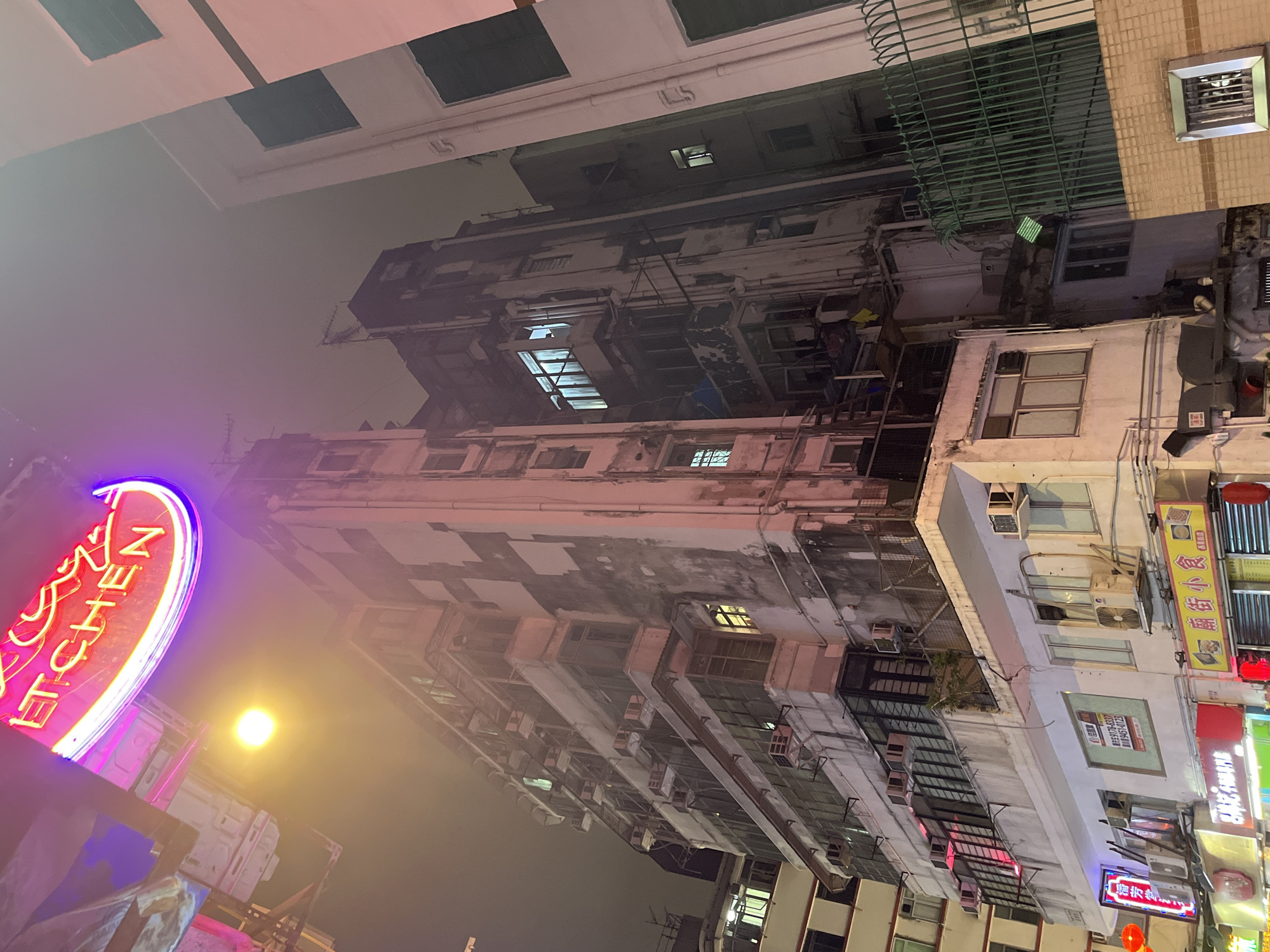 The image depicts a nighttime street scene in Hong Kong, China, characterized by tall, densely packed buildings. The architecture is worn and aged, with visible signs of weathering and disrepair on the facades. A prominent neon sign in pink and purple colors is visible on the left side of the frame, indicating a business establishment, possibly a restaurant. Street lighting casts a bright glow, and various smaller signs in Chinese characters are visible on the building exteriors. Air conditioning units are affixed to the buildings, indicating a humid climate. The sky is hazy, contributing to the overall gritty atmosphere.