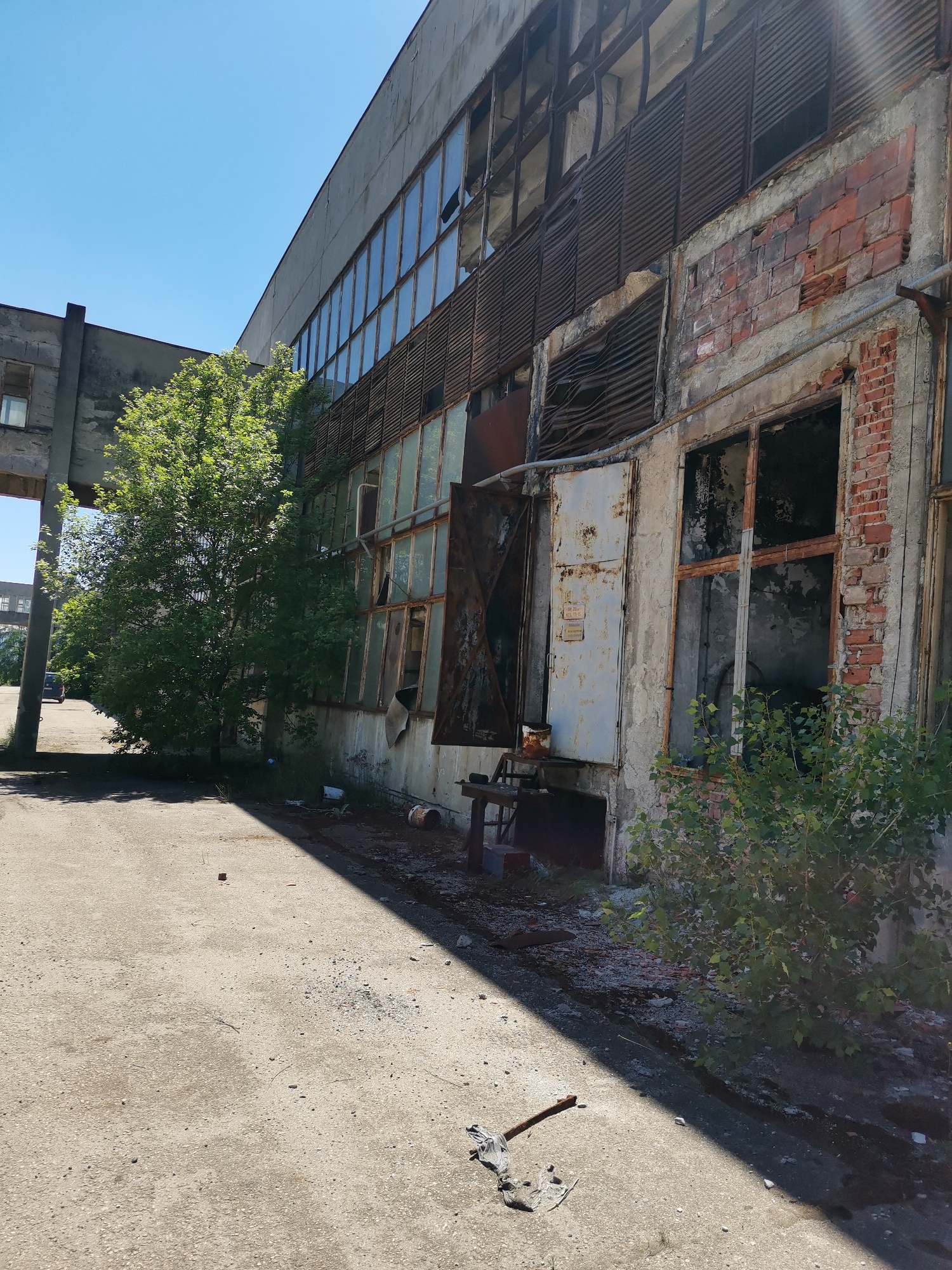 The image shows a deteriorated, abandoned building in what is purportedly the city of Verinsko, Bulgaria. The structure is a long, industrial-style building with a combination of concrete, brick, and rusting metal elements. Many of the windows are broken or missing. Some appear to be boarded up. The ground is cracked asphalt. Debris is scattered near the building, including what looks like broken bricks. A tree grows near the structure. An axe and debris are in the foreground.