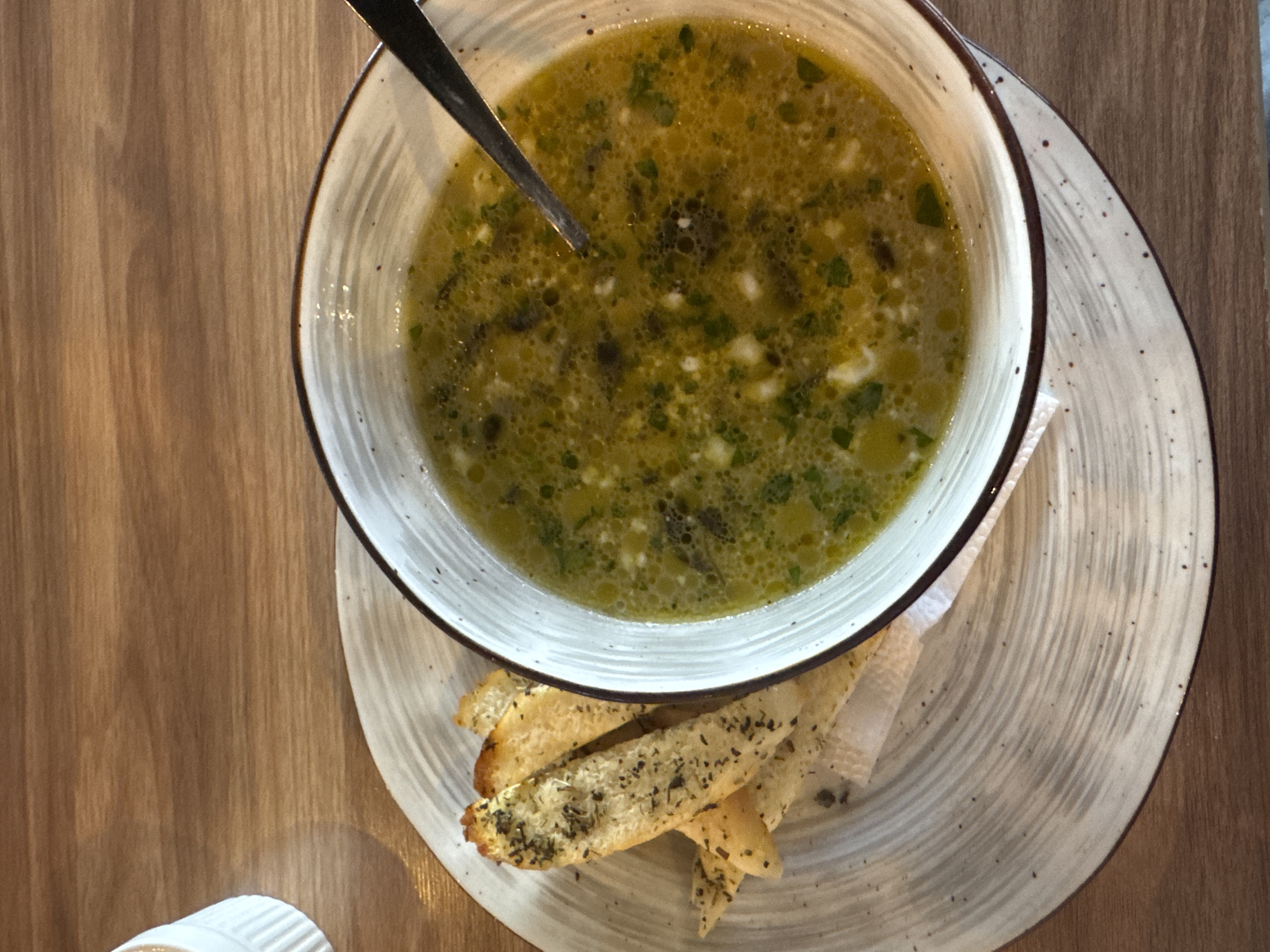 A top-down shot captures a bowl of green soup served with toasted breadsticks. The soup, a light green broth with visible flecks of herbs and some small white pieces, is presented in a rustic, cream-colored bowl with a brown rim. A dark metal spoon rests in the soup, angled towards the left. The bowl sits on a circular, speckled plate with a textured, grey and white striped pattern. Beneath the plate and to the left of the bowl are several golden-brown breadsticks, sprinkled with dried herbs. The entire setting is placed on a wooden surface with a visible grain pattern. A small, white ridged object, possibly a salt shaker, is partially visible in the bottom left corner. The lighting suggests an indoor setting, possibly a restaurant. The scene evokes a sense of a meal being served.