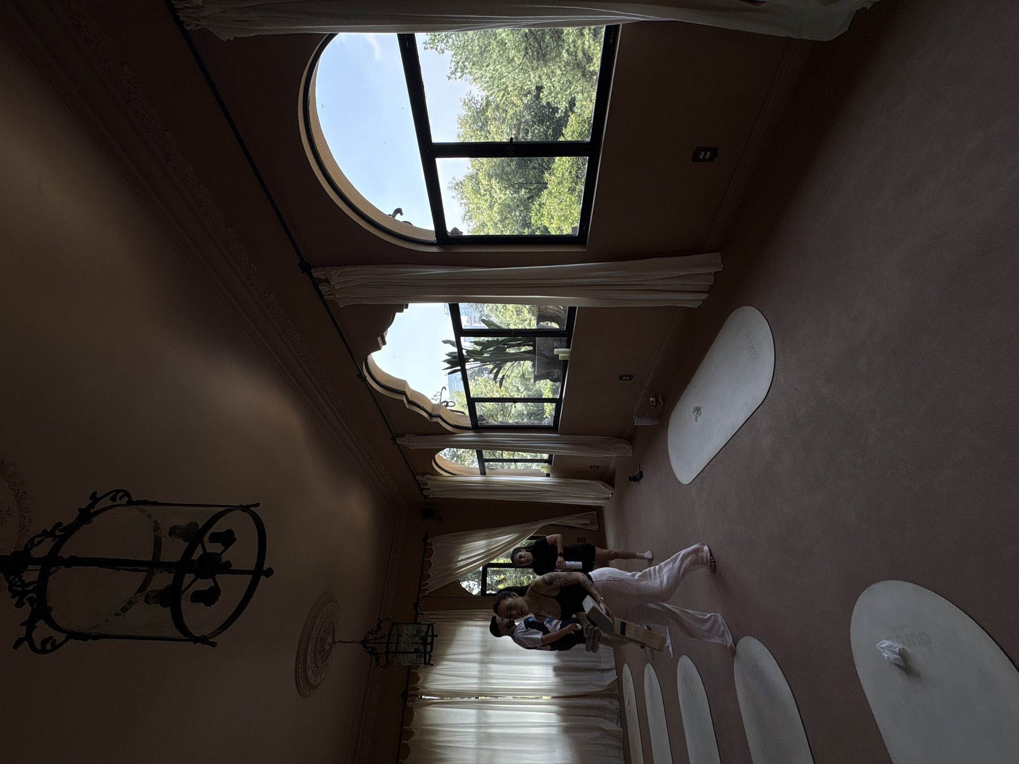 The image depicts an indoor yoga studio with large arched windows overlooking a densely wooded area, suggesting a location within or near a large park in Mexico City. The room has a warm, earthy-toned color palette with pink walls and a dark brown carpeted floor. A large, ornate chandelier hangs from the ceiling, adding a touch of vintage elegance. Several yoga mats are laid out on the floor, indicating that a class is in session or has recently concluded.
In the foreground, a woman with tattoos on her arm is walking towards the viewer, carrying yoga props. She is dressed in loose-fitting light pink pants and a dark tank top. Behind her, a man and a woman are standing, and another woman is in the background. The people appear to be participants or instructors.
The windows reveal a lush green canopy of trees, characteristic of the expansive green spaces found in Mexico City, such as Bosque de Chapultepec. The arched architecture of the windows, along with the decorative molding on the walls and ceiling, suggests a building with some historical or architectural significance. The lighting in the room is a combination of natural light filtering through the windows and the dim illumination from the chandelier.