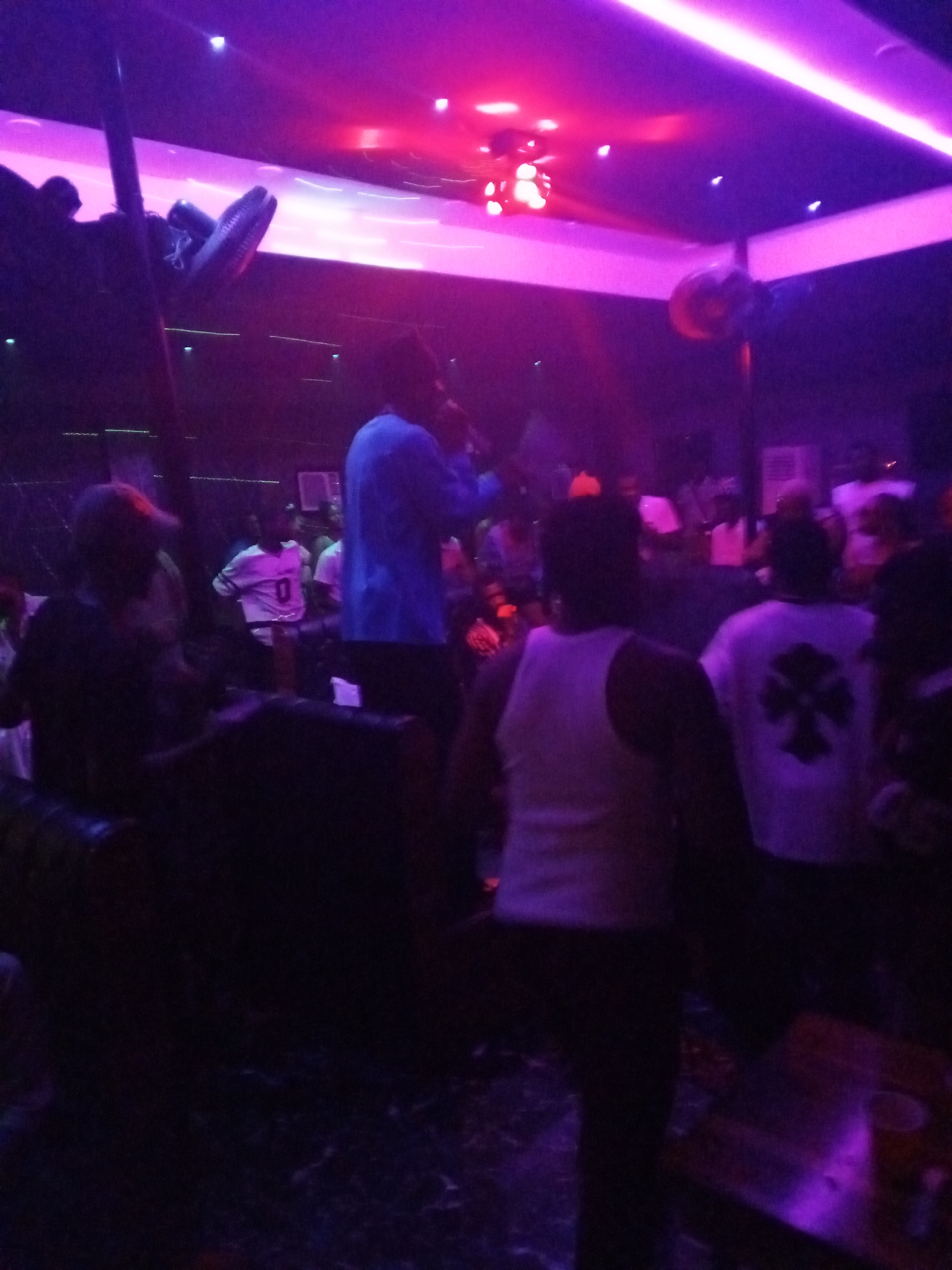 A nightclub scene in Unknown, Unknown, showing a crowd of people. Fluorescent lighting casts a pink and purple glow. Several people are standing, while others are seated. The background is dark and details are difficult to discern. A ceiling fan is visible. People's clothing includes white tank tops, t-shirts with designs, and a blue shirt.