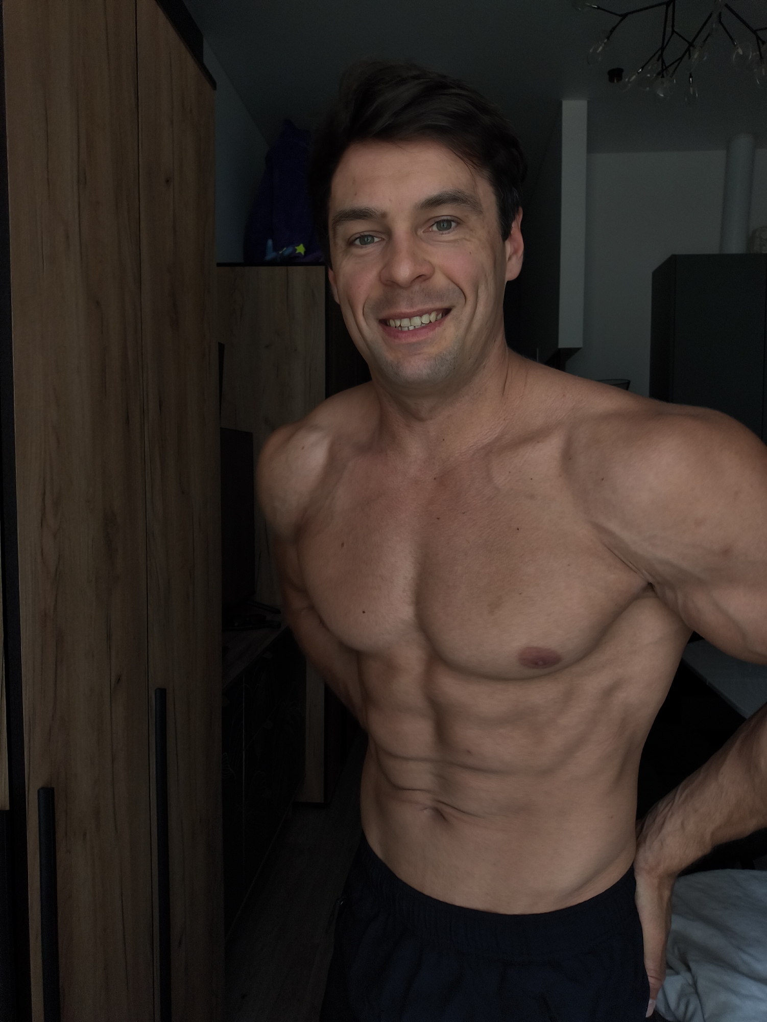 A man with dark hair and green eyes is standing in front of a wooden wardrobe. He is shirtless, revealing a muscular physique with well-defined abs and pecs. He is wearing black shorts. The man is smiling and looking towards the camera. The background shows a dark room with some furniture and what appears to be a closet. The scene is indoors, likely in a residential setting.