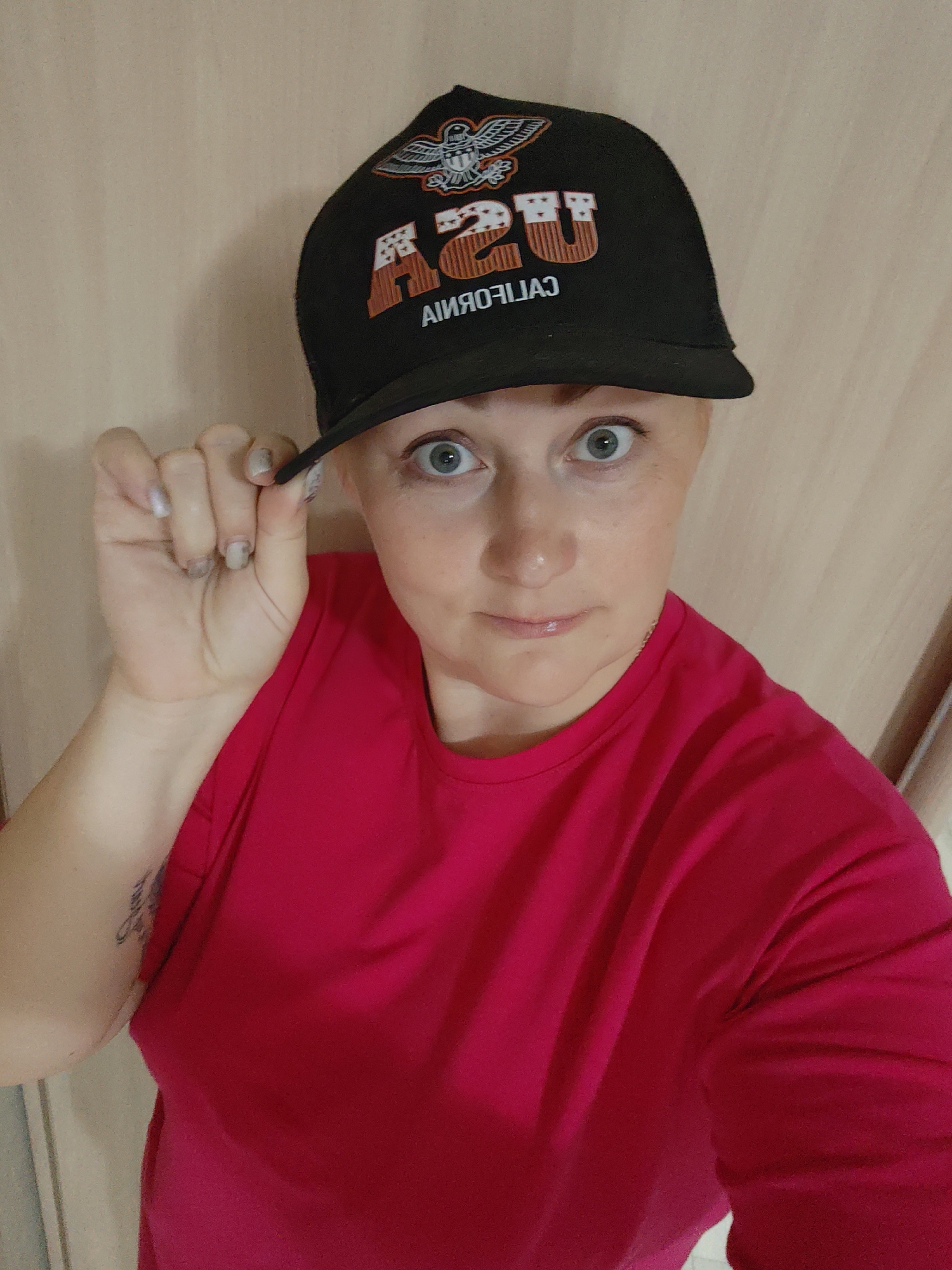 A woman with short, light-colored hair and pale skin is pictured in a selfie. She is wearing a bright red shirt and a black baseball cap with a graphic design featuring "USA" and "CALIFORNIA" in reverse writing. Her left hand is lifting the brim of the cap. Her eyes are wide and appear light blue. A tattoo is visible on her left arm. The background is a light, neutral-toned wall with vertical panels. Given the request of including the location Марковский, Russia, without any indication the image was taken there, I can only speculate the image may be taken indoors in a house or building somewhere in that city.