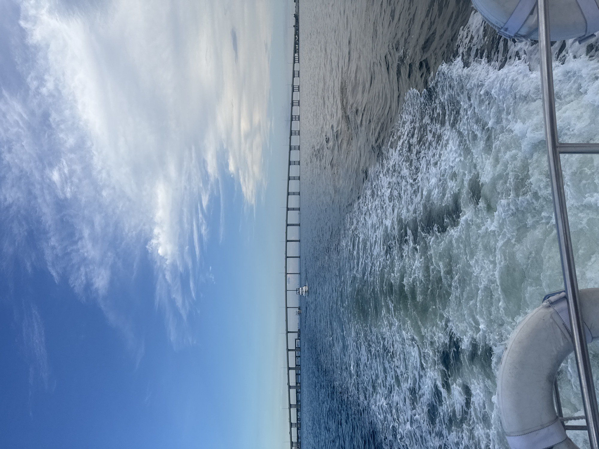 The image shows a wide view of a body of water, likely a bay or strait, with a long bridge stretching across the horizon. The perspective is from a moving boat, indicated by the frothy white wake trailing behind. The water is a deep blue, with the wake creating a textured contrast. The sky above is a mixture of clear blue and scattered white clouds, with a distinct line where the clouds become denser and lighter. Two white boats are visible on the water in the distance, approaching the bridge. The bridge itself is a multi-lane structure supported by numerous concrete pillars, and vehicles can be seen on its surface. The scene captures a moment of transit on the water, with the vast expanse of the sky and sea dominating the view, and the man-made structure of the bridge providing a sense of scale and human presence. This appears to be the Rickenbacker Causeway in Miami, Florida, given the characteristic long bridge over Biscayne Bay.