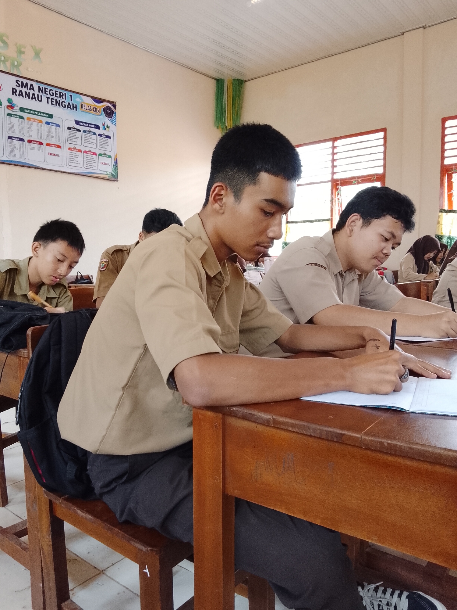 In a classroom at SMA Negeri 1 Ranau Tengah, located in Tanjung Setia, Indonesia, students in tan uniforms are seated at wooden desks, engaged in writing. The foreground features two male students actively participating in a writing activity, one in sharp focus and the other slightly blurred. The student in sharp focus, on the right, is leaning over a notebook, pen in hand, with his arm resting on the desk. Behind him, another student also appears to be writing. To the left, a third student is depicted with his head down, looking at his desk. The classroom environment is suggested by the presence of desks and a notice board on the wall displaying class schedules and other information. The lighting appears to be natural, coming from windows with horizontal blinds. The overall scene depicts a typical classroom setting during a lesson or examination.