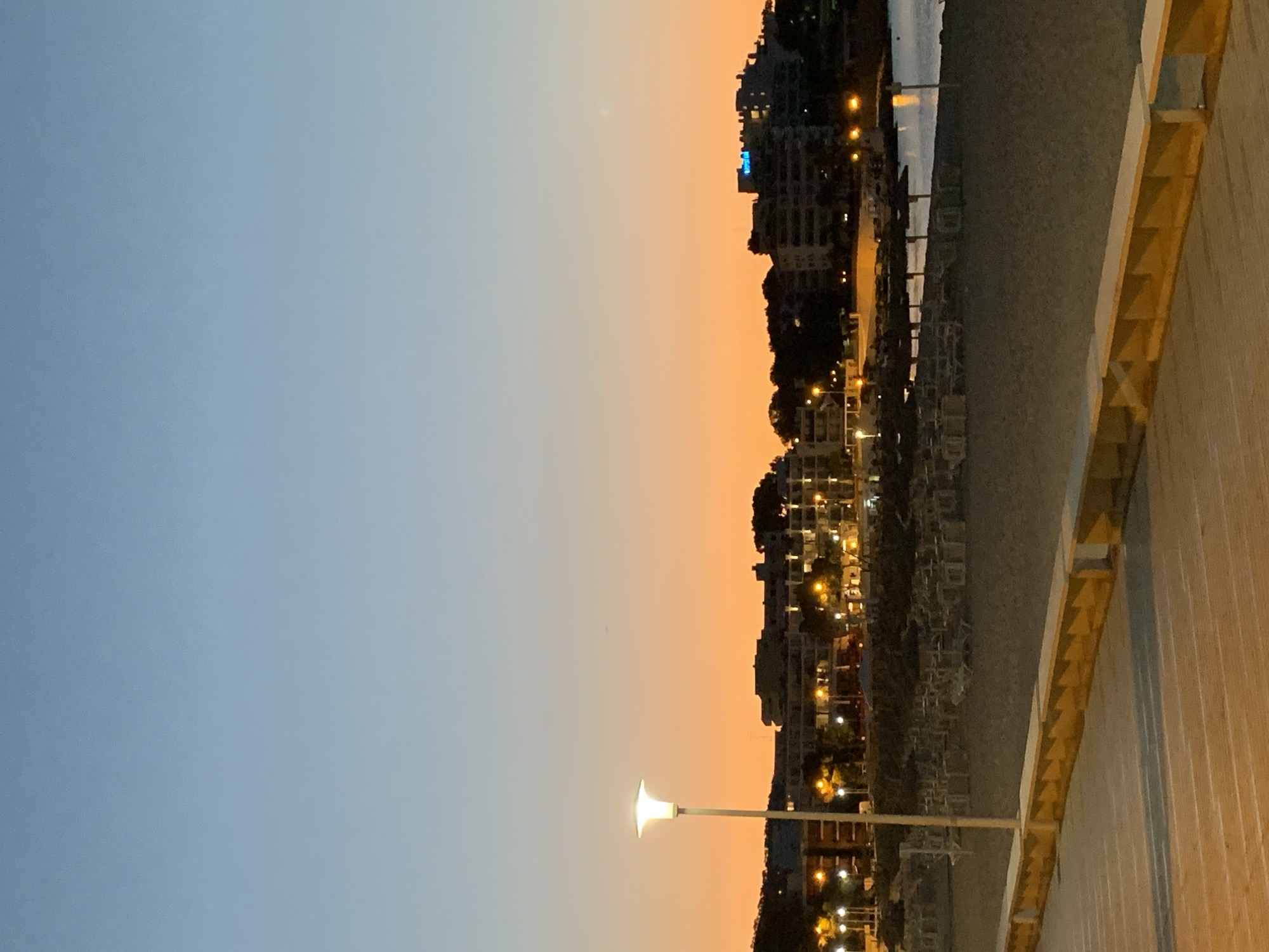 The image depicts a beach at dusk in Palmanova, Spain. The sky transitions from a pale blue at the top to a vibrant orange near the horizon, indicating either sunrise or sunset. In the background, a town is visible with several buildings, many of which are illuminated with artificial lights. A prominent streetlamp stands tall on the left side of the frame, casting a bright glow. The beach itself is sparsely populated, with rows of empty lounge chairs and umbrellas suggesting it was recently in use. A wooden boardwalk or promenade runs along the foreground, with a textured surface and a yellow-capped railing. The sea is calm and reflects the lights of the town. No people are visible in the image.