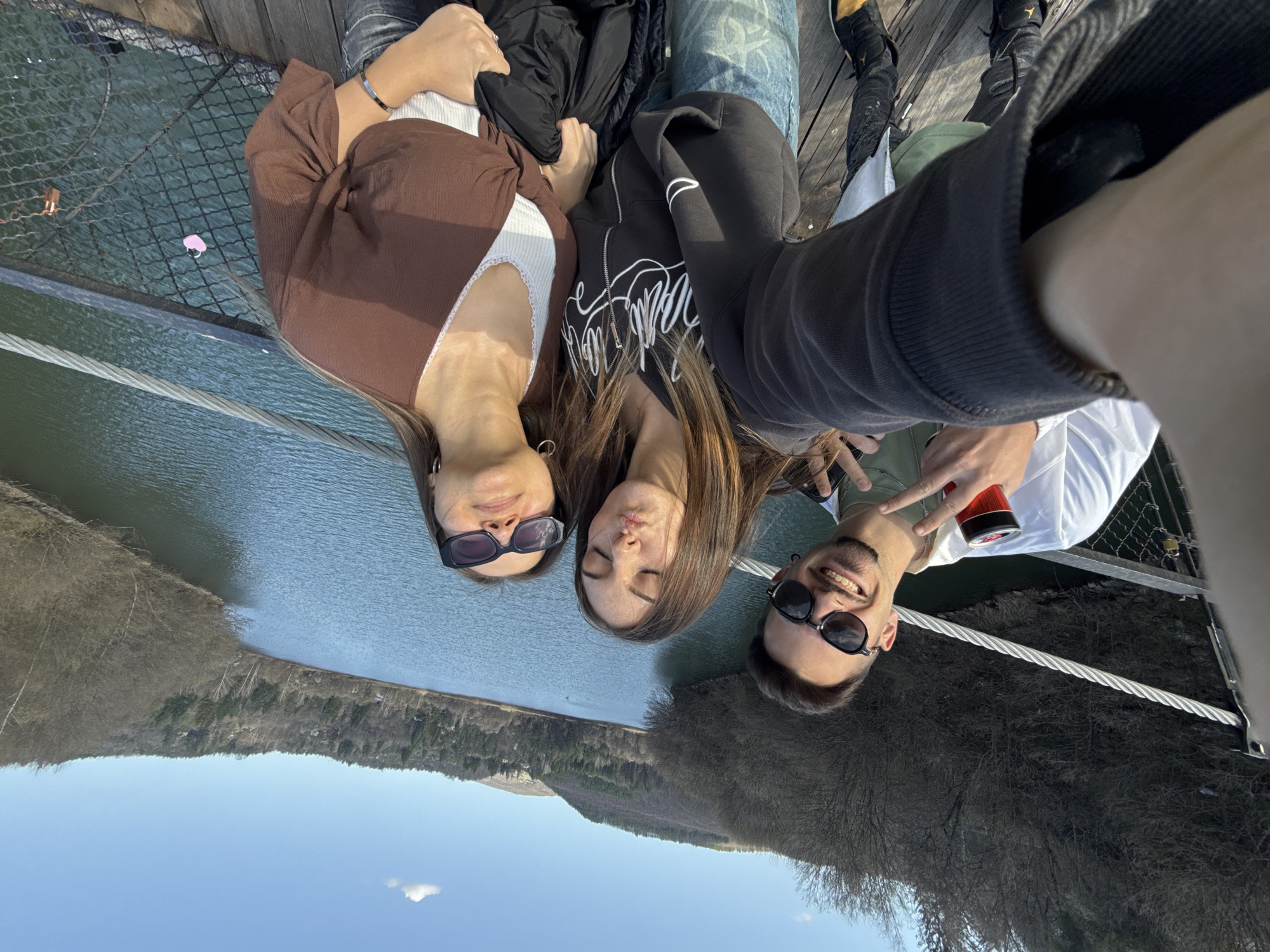 Three people, two women and one man, are posing for a selfie while standing on a bridge. The location is likely near Dolni Pasarel, Bulgaria, given the landscape featuring a reservoir and forested hills. The man is holding a red can in his left hand. The bridge has metal railings. The sky is visible in the background.