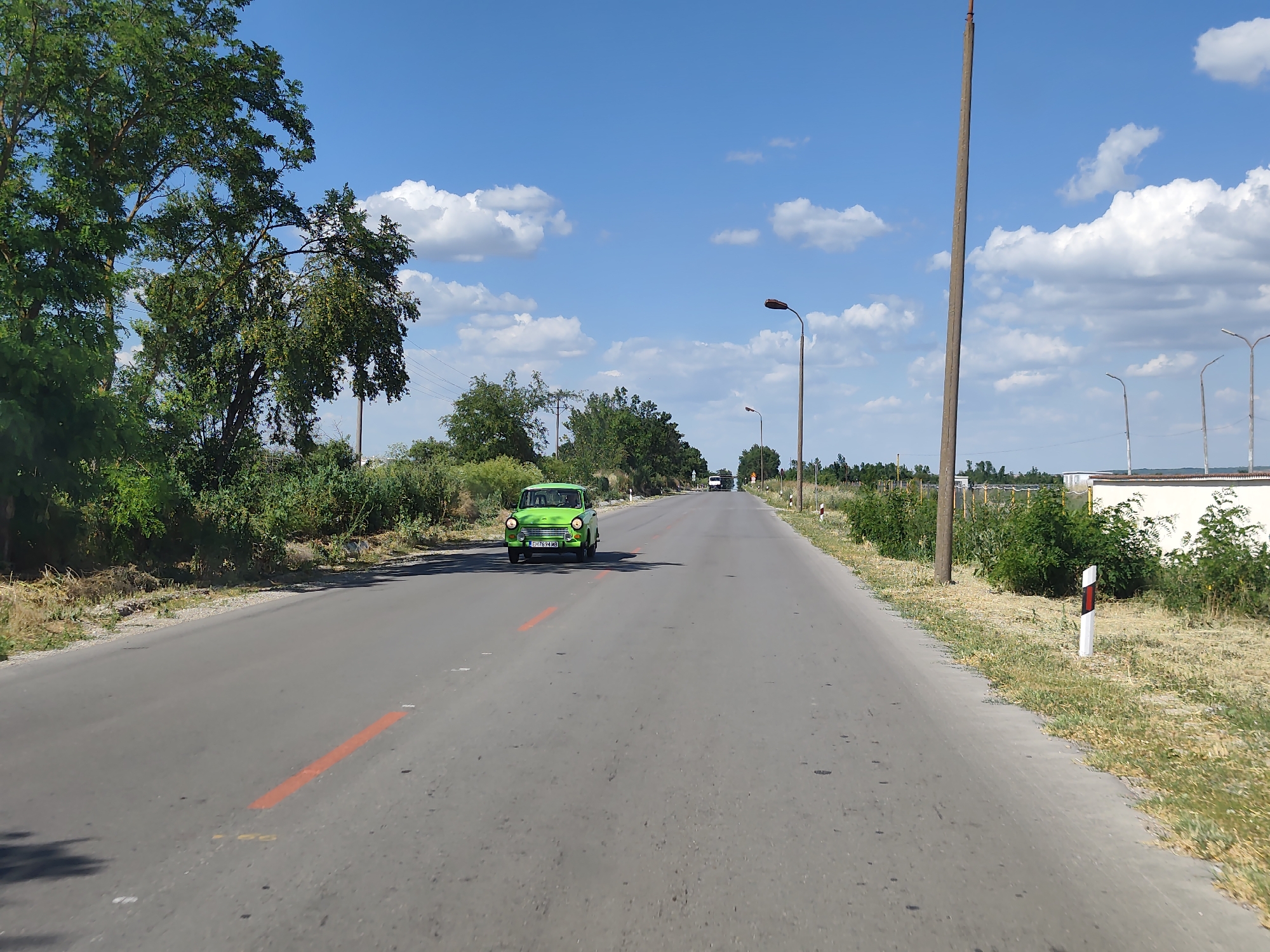 A bright green, vintage car is driving down a paved road. The car is centrally located in the frame, driving towards the viewer on the road. The road has orange markings on it. On either side of the road are green trees and bushes, with dry, yellow grass. In the background, there are a few more trees and what appears to be a white building with a fence. The sky is blue with scattered white clouds. There are several utility poles visible along the right side of the road, some with streetlights. One utility pole on the right is very tall and rustic. There are also road markers on the right side of the road with red and white stripes. The scene appears to be set in a rural or suburban area. The time of day appears to be daytime, likely mid-morning or afternoon, given the bright sunlight and shadows. The weather appears to be clear and sunny. The license plate on the car reads "CH 7624 MS".