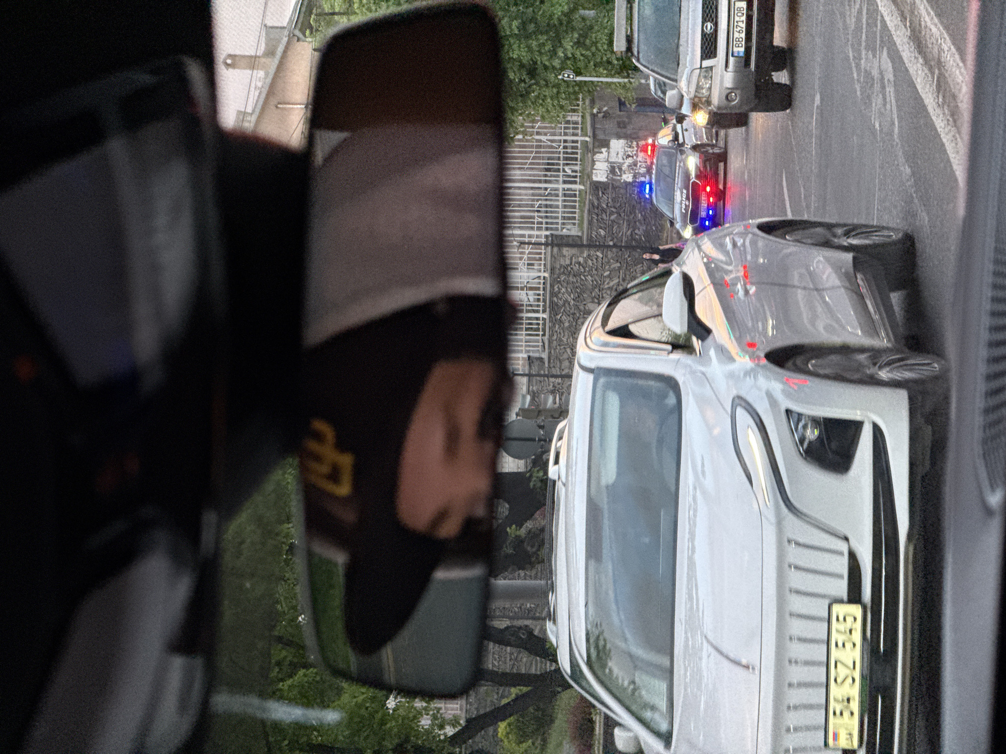 The image depicts a street scene in Yerevan, Armenia, as viewed from inside a vehicle. A person's face is reflected in the car's side mirror. Outside, several vehicles are visible, including a silver SUV with the license plate "BB 671 QB", a police car with flashing red and blue lights, and a light-colored vehicle with the license plate "54 SZ 545". The street is lined with trees and buildings, and a wrought-iron fence is partially visible. A person is standing next to the police car.