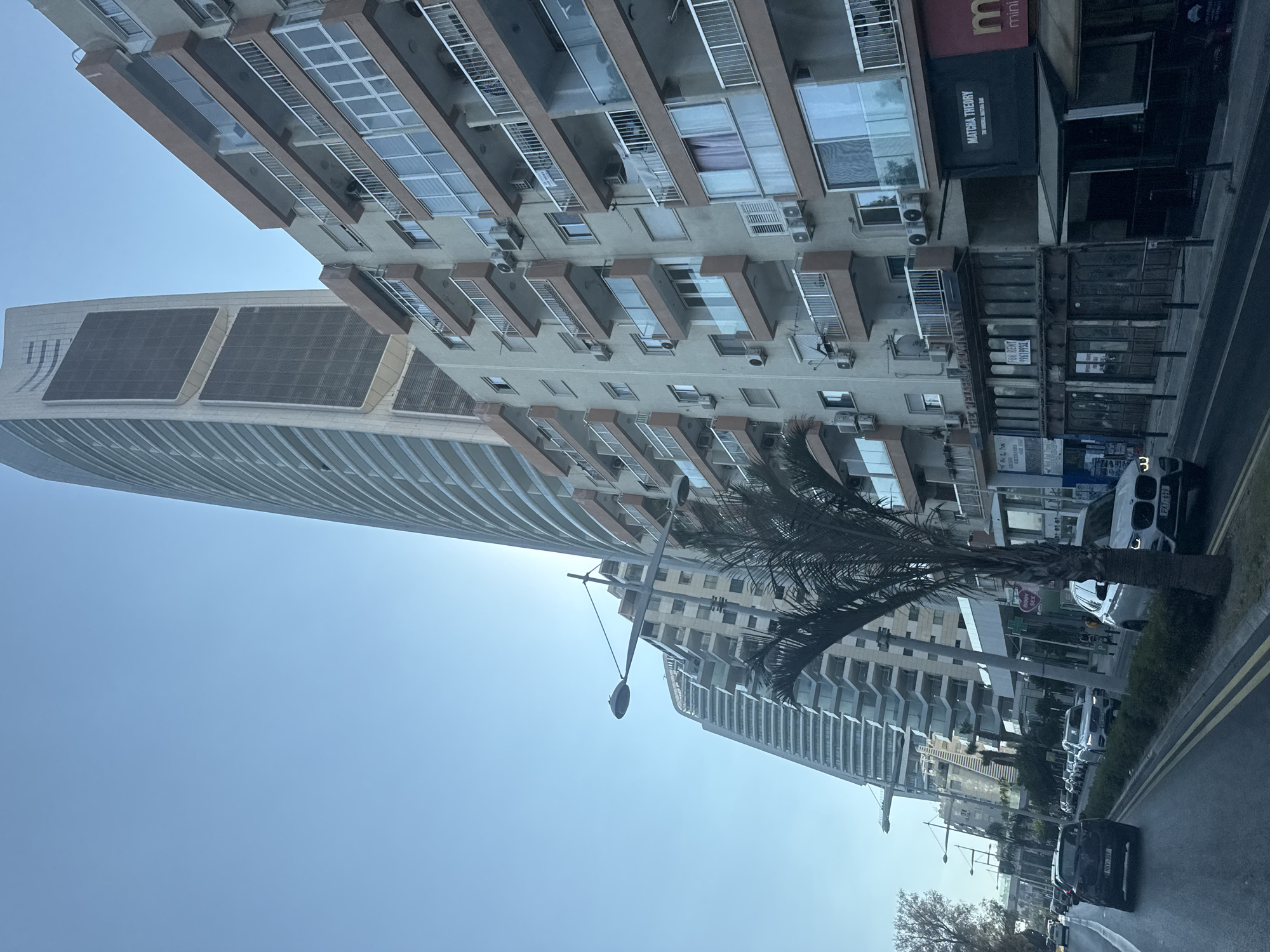 A street view in Limassol, Cyprus, shows a modern cityscape with a clear blue sky. In the foreground, a street with traffic is visible, including a black BMW SUV and a white BMW SUV. On the right side of the street, there are buildings with balconies, and one prominent tall, curved skyscraper with a dark, patterned facade dominates the background. A palm tree is partially obstructing the view of the buildings. Various shops and businesses are located on the ground floor of the buildings, including "MATCHA THEORY" and "mini". The image appears to be taken from a moving vehicle.