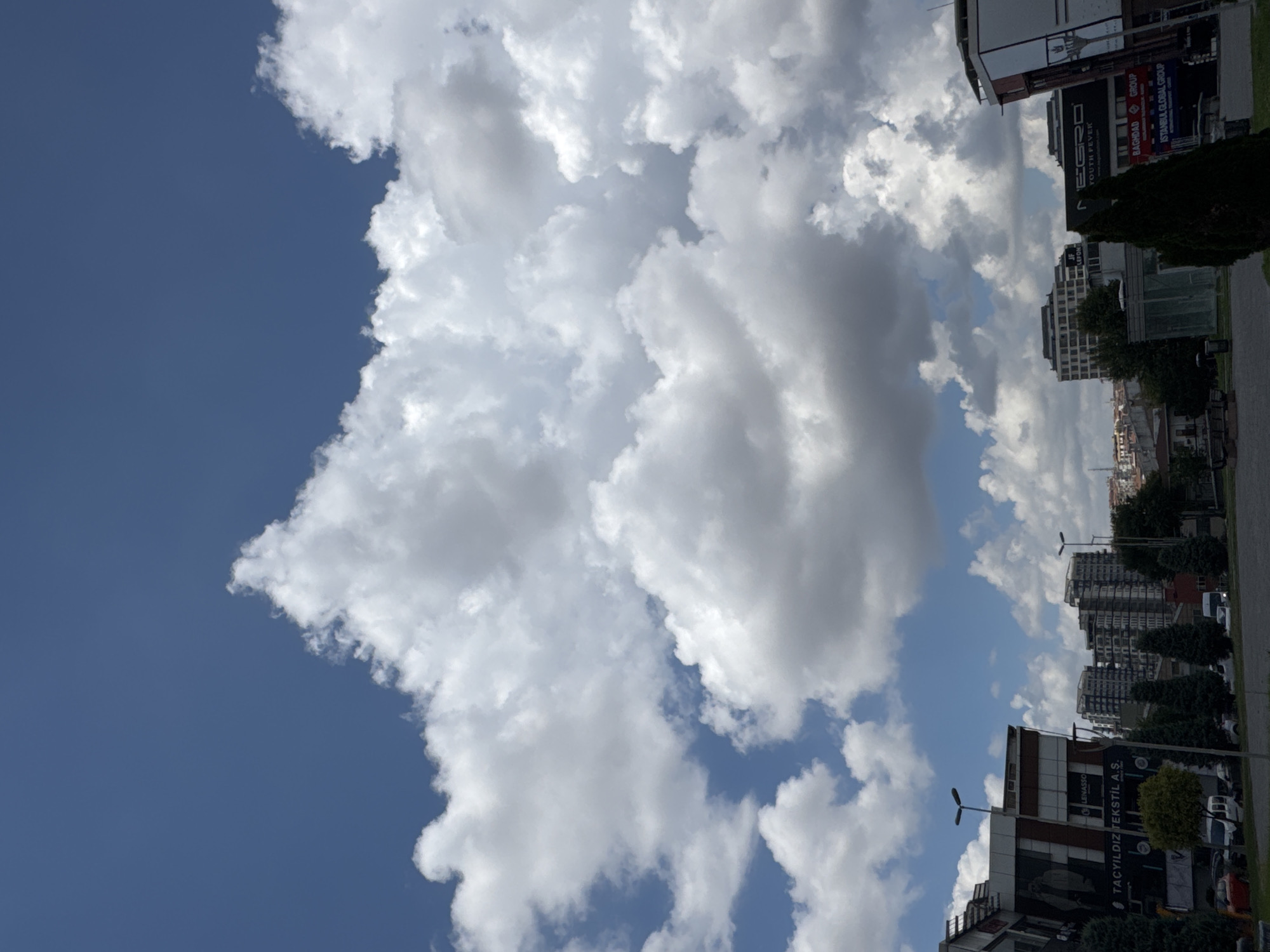 In the city of Gungoren, Turkey, this image captures a view of buildings and a street scene under a cloudy blue sky. The foreground features a paved area with several trees, including cypress trees, and some park benches. Beyond this, a street is visible with parked vehicles, including vans and cars. On the left side of the image, a modern building with large windows and signage that reads "TACYILDIZ TEKSTIL A.S." is prominent. Further back, several multi-story residential and commercial buildings dominate the skyline, some with visible balconies and architectural details. The sky is filled with dramatic white cumulus clouds against a clear blue backdrop, suggesting fair weather with cloud cover. The overall scene is one of urban development within a bustling Turkish city.