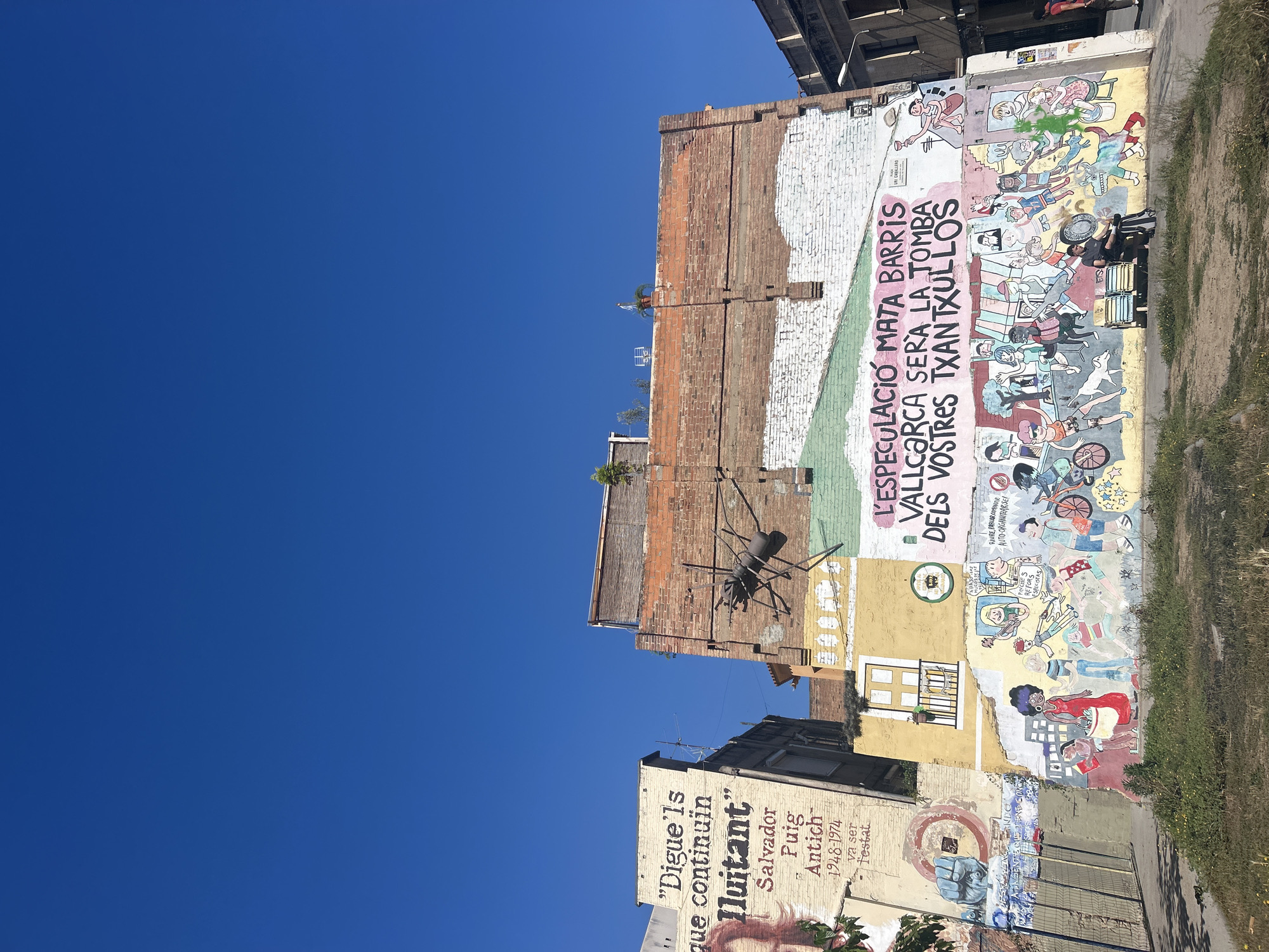 The image shows a colorful mural on the side of a building in Barcelona, Spain. The mural features a variety of cartoonish characters and political slogans in Catalan. A large metal sculpture of a spider is affixed to the building above the mural. The sky is bright blue, and part of another building with a mural is visible below. The mural reads: "L'ESPECULACIÓ MATA BARRIS, VALLCARCA SERÀ LA TOMBA DELS VOSTRES TXANTXULLOS" or "Speculation Kills Neighborhoods, Vallcarca Will Be the Tomb of Your Deals." Another portion of the mural reads "Digue'ls que continuïn lluitant" Salvador Puig Antich 1948-1974 va ser testat", or "Tell them to keep fighting! Salvador Puig Antich 1948-1974 was tested".