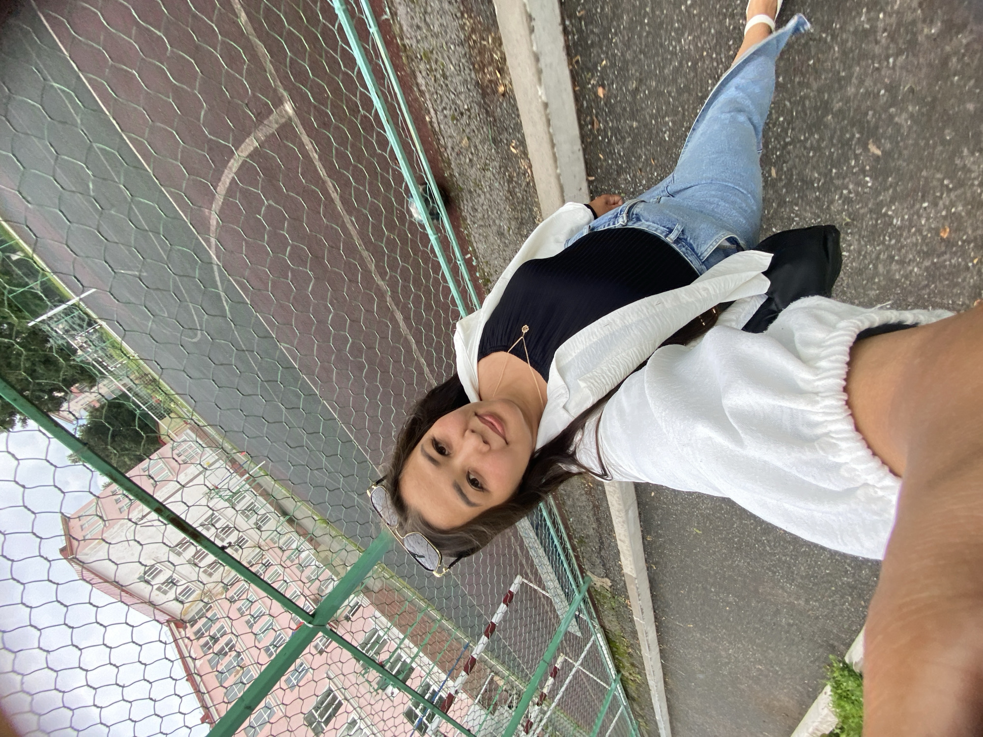 A young woman takes a selfie on an outdoor sports court, possibly in Ufa, Russia. The court, with its reddish-brown surface and white markings, is enclosed by a green wire mesh fence. In the background, a large, pink, multi-story building with numerous windows is visible, alongside trees and a cloudy sky. The woman, wearing a black top, blue jeans, a white open shirt, and sunglasses on her head, is looking at the camera and smiling. She has a small necklace with a pendant. The perspective suggests she is standing and holding the camera at a high angle. The ground around the court is asphalt.