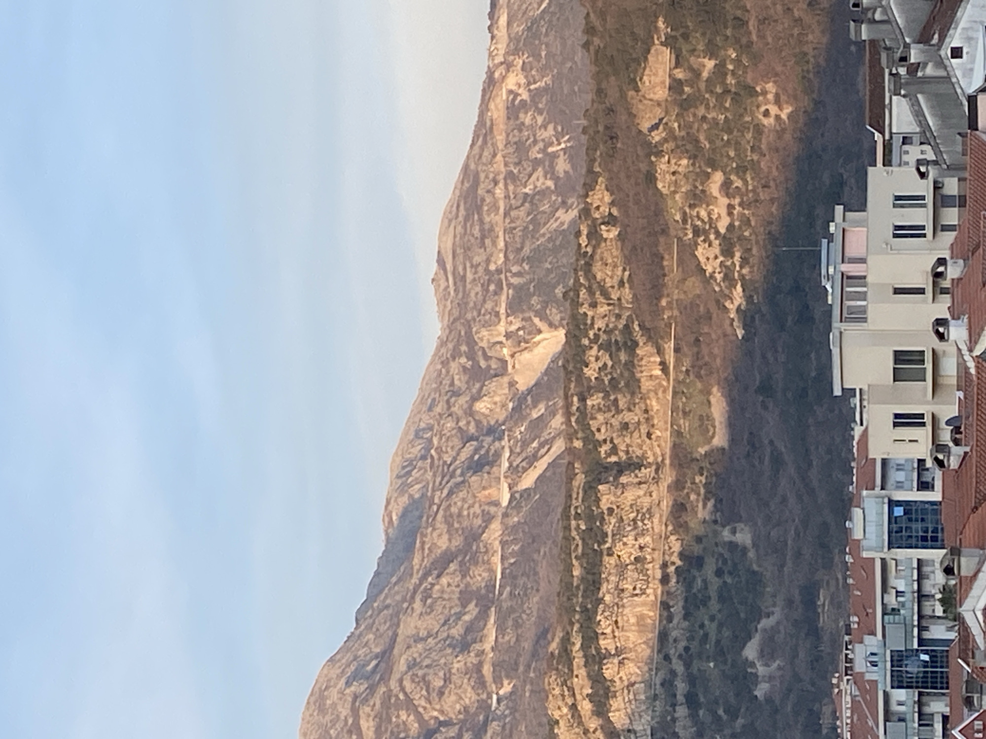 The image captures a panoramic view of Budva, Montenegro, with a prominent mountain range dominating the background. The upper two-thirds of the frame feature rugged, multi-layered mountains, displaying a mix of exposed rock formations and sparse vegetation. Horizontal cut roads or tracks are visible traversing the mountain slopes at various elevations. The upper and middle sections of the mountains are illuminated by warm, golden sunlight, contrasting with the darker, shadowed lower slopes which exhibit denser, darker green and brown vegetation. The sky above is clear and light blue. The lower third of the image depicts an urban residential area of Budva, consisting of numerous multi-story buildings predominantly featuring terracotta-colored tiled roofs. Some buildings present light-colored facades and modern architectural elements, including blue-tinted glass window panels. No individuals, vehicles, or specific activities are discernible within the frame.