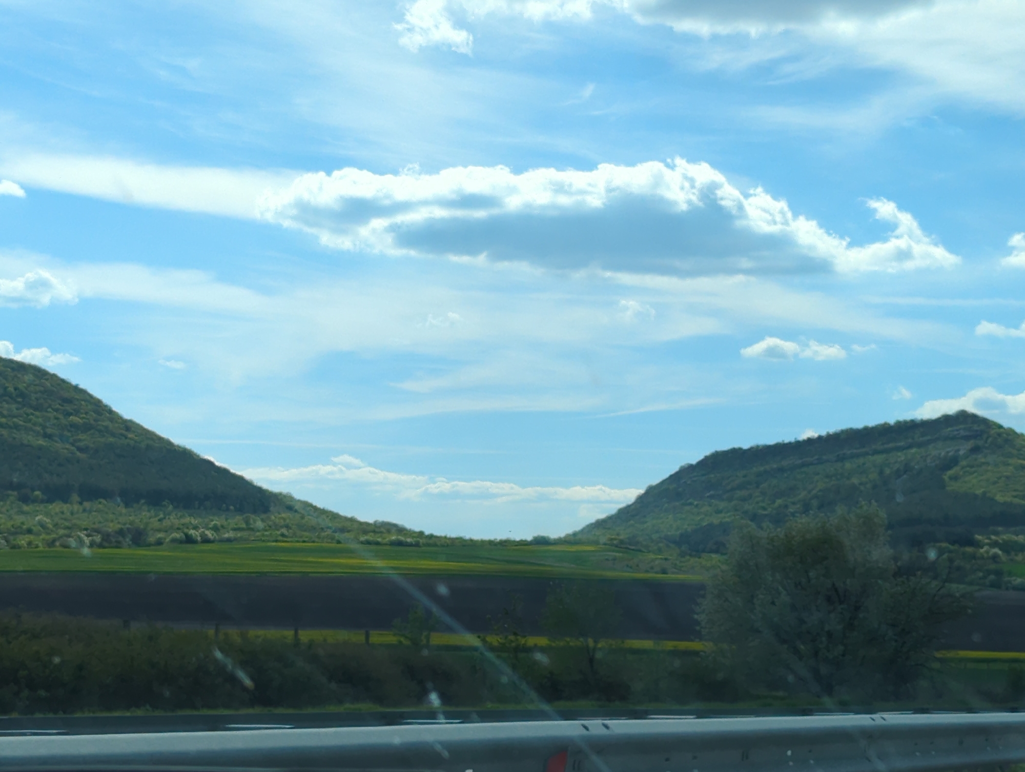 This image presents a serene daytime landscape observed from what appears to be a moving vehicle near Nevsha, Bulgaria. The lower part of the frame features a metallic silver-grey guardrail, indicating the perspective is from a road or highway.
The sky dominates the upper half, a vibrant blue canvas adorned with various white clouds. Fluffy cumulus clouds are scattered across the mid-sky, while delicate, wispy cirrus clouds stretch across the upper regions. Sunlight brightly illuminates these clouds, creating a sense of depth and a cheerful atmosphere.
Below the expansive sky, two verdant hills or mountains frame the central vista. Their slopes are densely covered in lush green trees and shrubs, with lighter green foliage and hints of blossoms suggesting late spring or early summer. Between these elevated landforms, the terrain flattens into agricultural fields. Some fields are a bright, almost neon green, possibly indicating young crops, while others appear as dark brown, recently tilled earth. A faint fence line is visible in the mid-ground on the left, separating different sections of the landscape. A solitary, light-foliaged tree, potentially a flowering species, stands on the right side of the road before the base of the hill.
The scene conveys a sense of peaceful natural beauty and wide-open spaces, characteristic of a rural European landscape. The bright light and clear skies indicate good weather conditions. No individuals or specific activities are discernible, focusing the attention purely on the scenic environment.