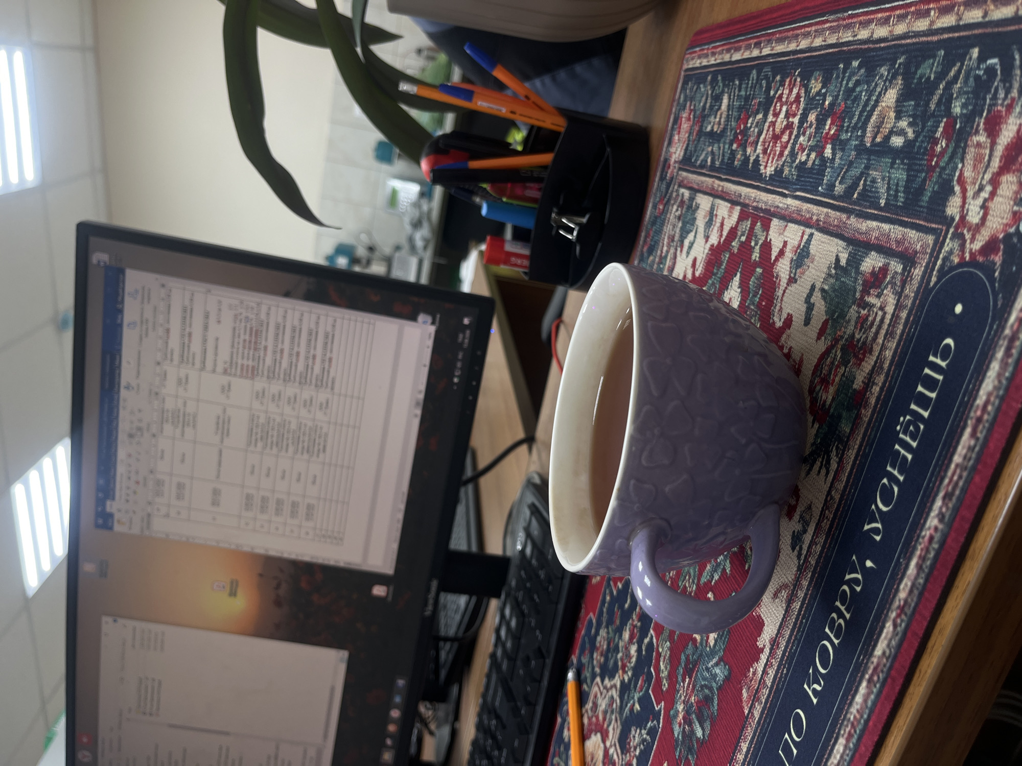 This is a close-up, eye-level shot of a desk with a cup of tea in the foreground. The cup is lavender with a textured pattern and sits on a decorative placemat with a red, blue, and beige oriental design. The placemat has text on it, possibly in Russian, with some visible letters including "ПО КОВРУ, УСЧЁШ". In the background, a computer monitor displays two windows, one showing a spreadsheet or table and the other a desktop wallpaper image of a sunset over a field. A black computer keyboard is visible in front of the monitor, and to the right of the monitor, there is a black pen holder filled with pens and pencils, including some orange ones. A hint of a green plant with large leaves is in the upper right corner, partially obscuring a beige object. The overall environment appears to be an office or study. There are no people visible in the image. The scene is well-lit, likely by overhead office lighting. The exact location in Rostov-on-Don, Russia cannot be determined from the visual information provided.