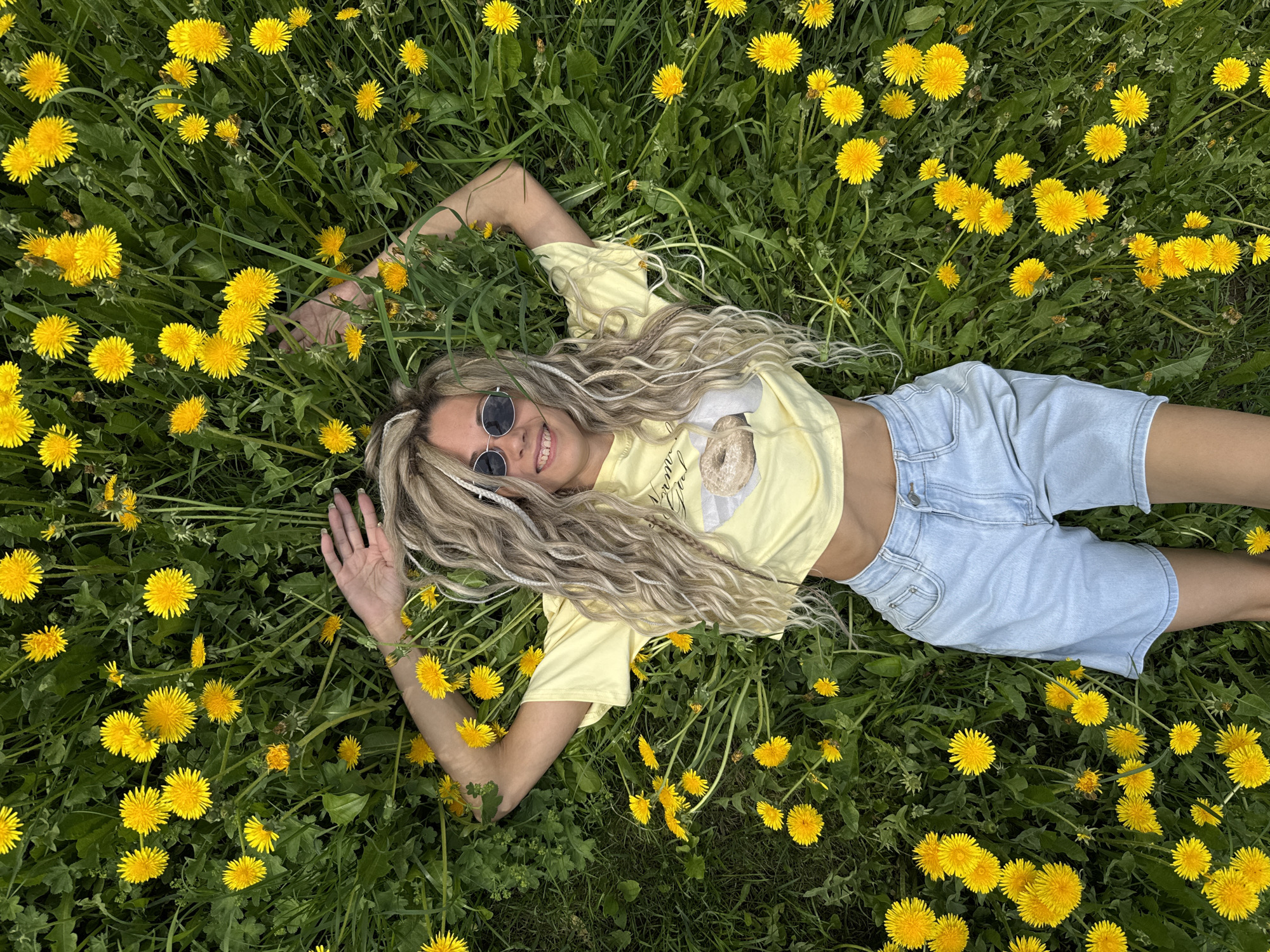 A woman lies on her back in a field of dandelions. She is wearing round sunglasses, a yellow t-shirt, and light blue denim shorts. Her arms are outstretched on either side of her, and she is smiling. The dandelions are in full bloom, and the grass is green. The location is Unknown, Unknown.