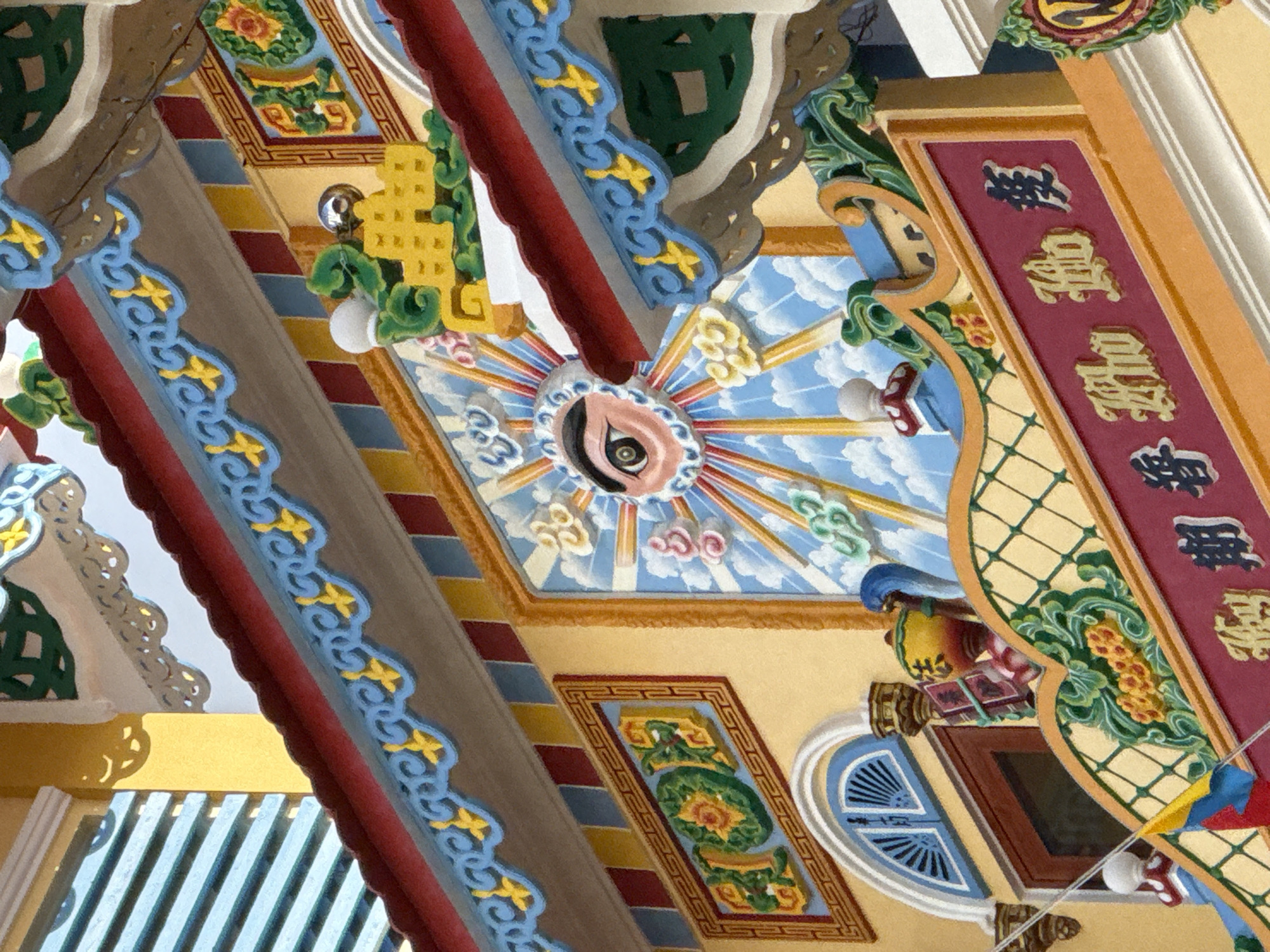 The image shows a close-up view of the ornate facade of a building, likely a temple or cultural site, in Nha Trang, Vietnam. The architecture is characterized by vibrant colors, intricate carvings, and decorative elements. A prominent feature is a large, stylized eye within a sunburst motif, painted against a backdrop of blue sky and white clouds. Surrounding this central artwork are decorative borders with blue, red, and yellow patterns, featuring floral and geometric designs. In the foreground, a deep red plaque displays gold-colored characters, likely Vietnamese or Chinese script, signifying an inscription or name. Other architectural details visible include a window with blue shutters, a decorative urn, and ornate roof eaves adorned with green and yellow motifs. The overall impression is one of rich cultural expression and detailed craftsmanship.