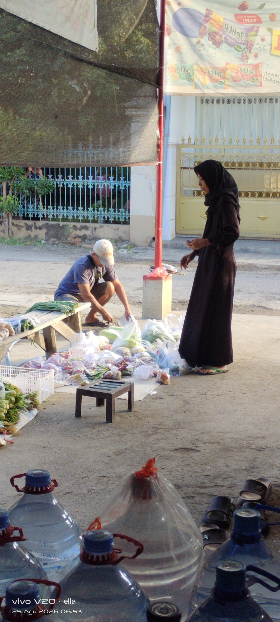 In Simo, Indonesia, an outdoor scene depicts a street-level market. Two individuals are present. A man wearing a blue t-shirt and a white baseball cap is crouched down near a wooden bench, sorting through produce. To his right, a woman wearing a dark hijab and a long dark dress stands near a red pole, holding something in her hands. A collection of vegetables and other goods, some in plastic bags and some in a white crate, are spread out on the ground and on the bench. Large water bottles are visible in the foreground. A small, dark wooden stool is placed in front of a pile of goods. In the background, a light-colored building with a yellow gate and banners advertising products are visible, along with a blue metal fence. The ground appears to be a dirt and asphalt mix.