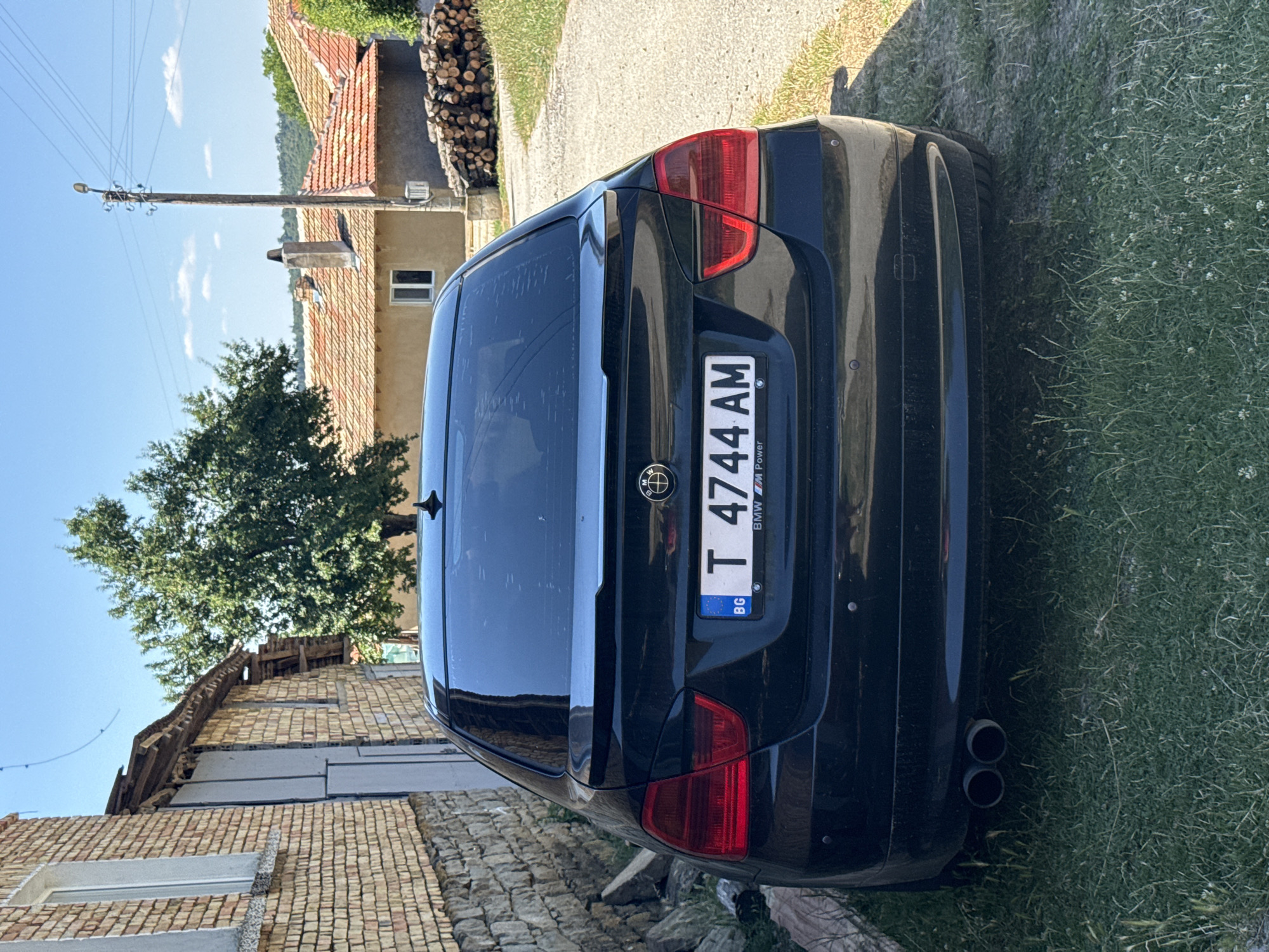 The image shows the rear of a black BMW sedan parked on a grassy area next to a brick building. The license plate reads "T 4744 AM" with a "BG" indicator. In the background, there are houses with tiled roofs, a wooden utility pole, and a stack of firewood. The sky is blue with some clouds. The location is identified as Konop, Bulgaria. There are no people visible in the image.