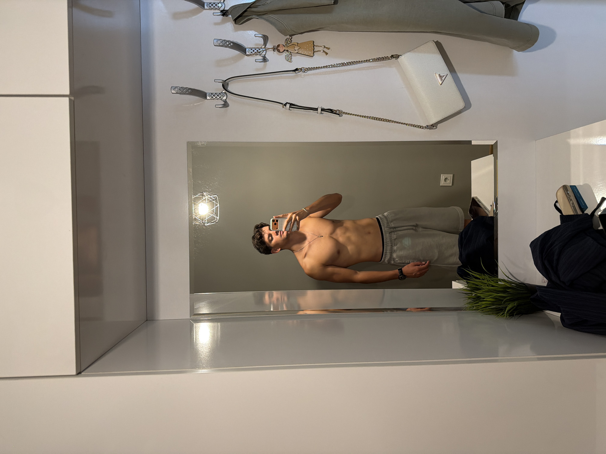 The image depicts a young man taking a mirror selfie in what appears to be an entryway or hallway. He is shirtless, wearing light gray athletic shorts, and a silver necklace. His reflection is visible in a large mirror. To the right of the mirror, several items are hanging on wall hooks, including a white purse with a gold chain, a light green jacket or vest, and a small angel charm. A floor lamp with a geometric shade hangs from the ceiling above the mirror. In the foreground, a small artificial plant in a black pot and some dark-colored fabric are visible on a white surface. The environment appears to be indoors, with modern white cabinetry and walls. The man's attire and the setting suggest a casual, personal space. The image does not contain any information that would suggest its location is in Blagoevgrad, Bulgaria.