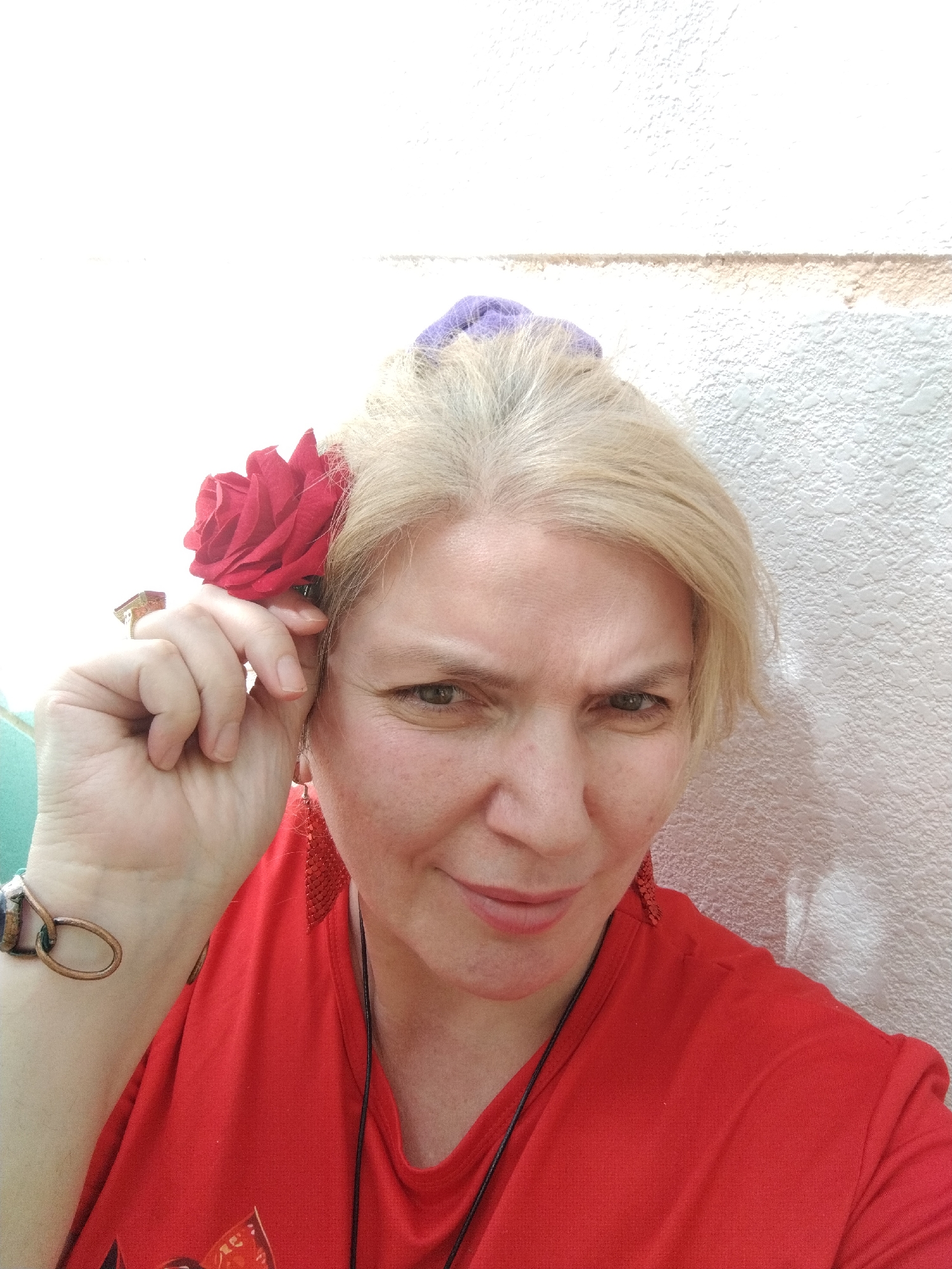 A mature woman is posing for a selfie against a light-colored wall, likely outdoors given the lighting. She is wearing a red t-shirt and has a red artificial rose tucked behind her ear on the left side of the frame. She has light blonde hair pulled back with a purple scrunchie, and is wearing dangling red earrings. She also has a ring on her left hand, and a bracelet on the same wrist. Her expression is slightly quizzical, with a slight smile and one eyebrow raised. There's no explicit indication in the image that places it in Al Mas'oudi, United Arab Emirates.