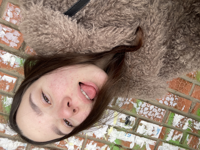 A young adult female is depicted in a close-up, slightly upward-angled shot against a brick wall, identified as occurring in the city of Ольховка, Russia. She has long, dark brown hair partially obscuring her right shoulder. Her face, characterized by dark eyebrows and light brown eyes, exhibits some redness and minor blemishes across the cheeks and forehead. She is looking directly towards the camera with her mouth open and tongue extended. She is wearing a textured, light brown or beige faux fur coat, with a black strap, possibly from a bag, visible over her right shoulder. The background consists of a reddish-brown brick wall, many bricks showing remnants of peeling white and green paint or plaster, and faint, indecipherable text or numbers on some surfaces.