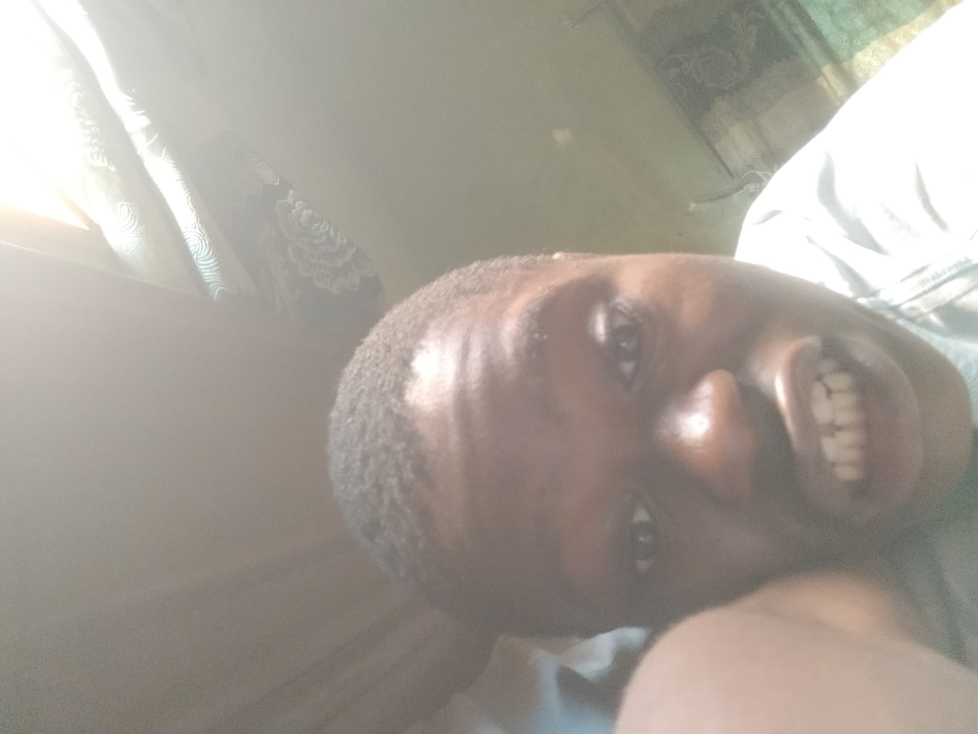 A close-up selfie of a man with dark skin. The background is blurry but appears to be an indoor setting, likely a bedroom, due to the presence of a window with curtains. The man is smiling broadly, showing his teeth. The image is overexposed, particularly on the left side near the window. The location is in Shangisha, Nigeria, though this cannot be verified from the image alone.