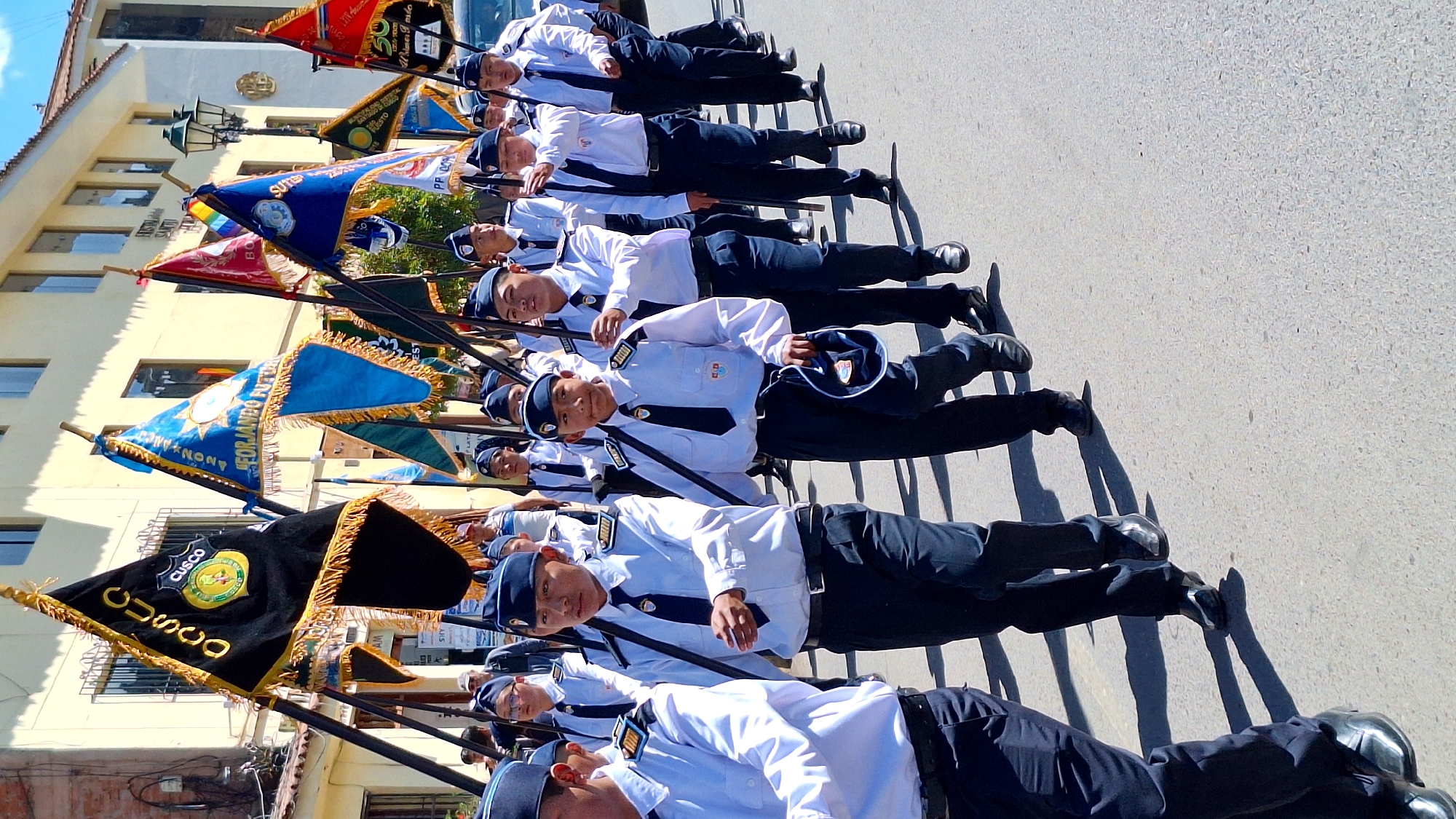 A parade is taking place on a sunlit street in Cusco, Peru, with a group of young people in uniform marching in formation. The individuals are wearing white collared shirts with dark ties and dark trousers, and blue caps. Many are holding poles with flags, including a large black flag with gold trim and the word "CUSCO" in gold lettering, and other flags in blue, red, and yellow. The buildings lining the street appear to be colonial in style, with yellow facades and arched windows. The ground is paved with concrete. The people are walking forward, suggesting a ceremonial procession.