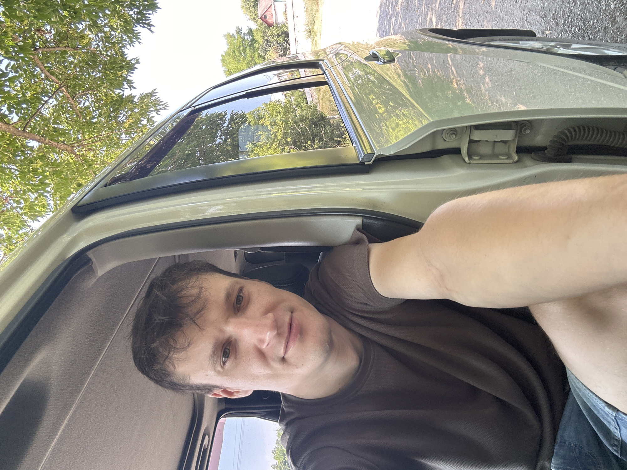 A young adult male is captured in a selfie from inside a light-colored vehicle, with the driver's side door open. He is positioned in the driver's seat, looking towards the camera with a slight smile. His right arm is extended out of the car, holding the phone for the selfie. He is wearing a dark brown t-shirt and denim shorts. The interior of the car is visible, including the roof lining and part of the dashboard. Outside the car, trees and a bright sky are visible, suggesting an outdoor setting during the daytime. The background shows a rural or suburban landscape with houses and foliage. The scene appears to be set in Makiv, Ukraine, given the context provided.