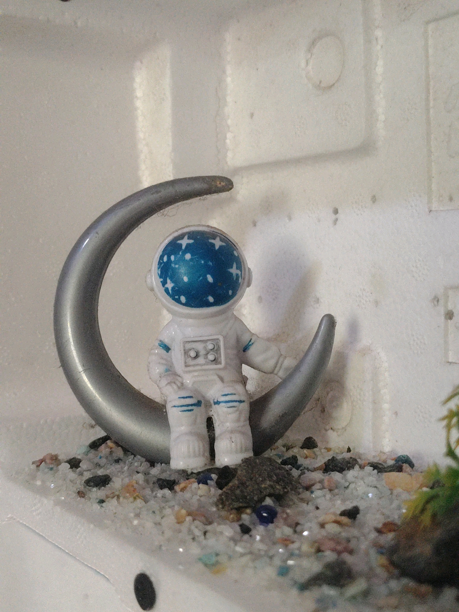 A close-up, eye-level shot shows a miniature astronaut figurine sitting on a crescent moon. The astronaut is white with blue accents on its suit and helmet, which is decorated with white stars. The moon is silver and curved, with the astronaut perched on its lower arc. The scene is set against a textured, white background. The ground beneath the moon is covered in a layer of small, multicolored pebbles and sand, with a few darker rocks scattered amongst them. In the lower right corner, a small, green leafy plant is partially visible. The lighting suggests an indoor setting, possibly a diorama or display. The overall impression is of a whimsical, space-themed scene. The image was taken in the city of Kinniya, Sri Lanka.