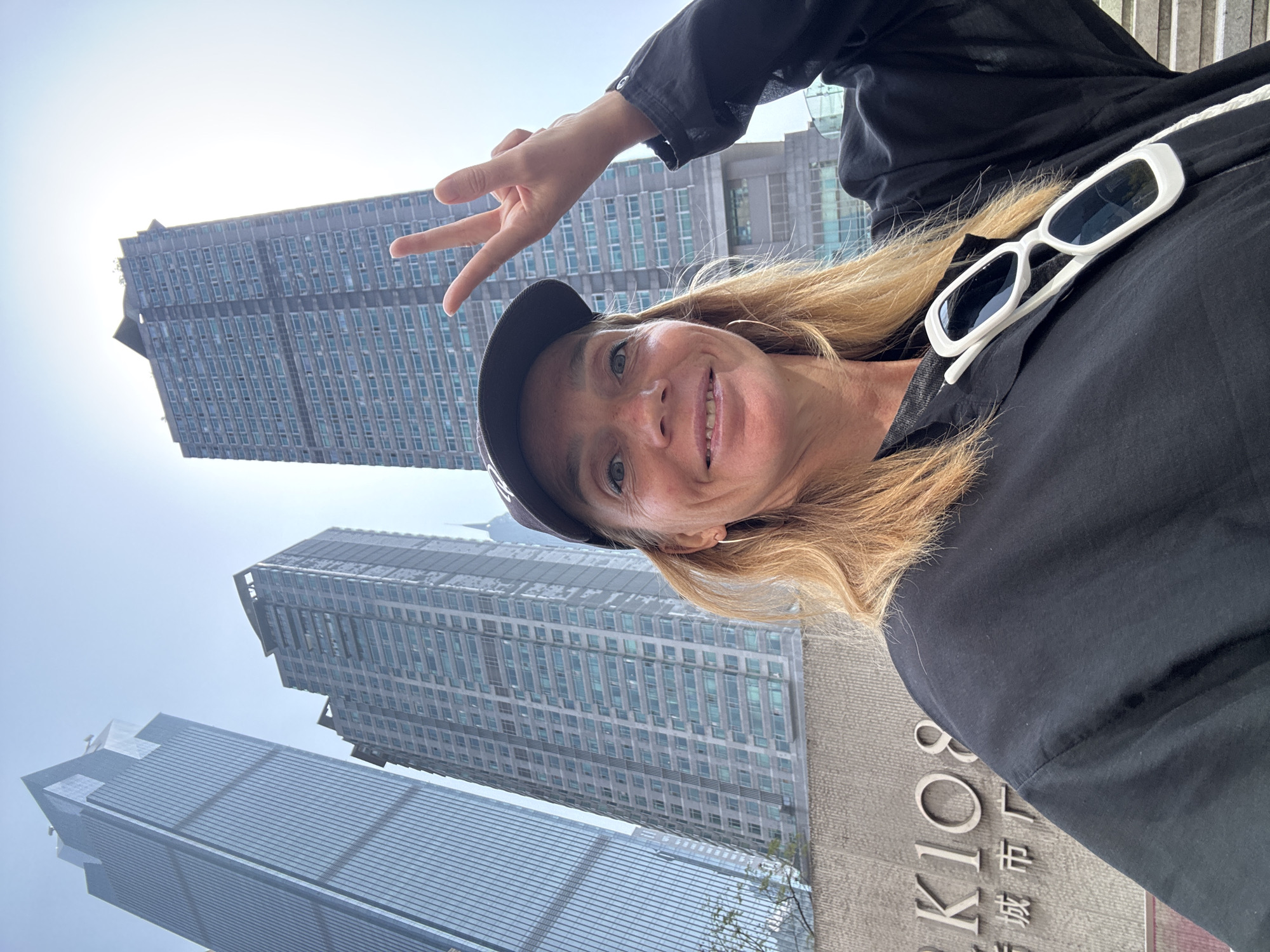 A woman is posing for a selfie in Yuzhong District, China. She is wearing a black baseball cap and a black shirt, and white sunglasses are tucked into the collar of her shirt. She is holding up a peace sign. Behind her are modern skyscrapers and a sign that reads "KI 108," likely indicating the location. The scene appears to be outdoors on a sunny day.