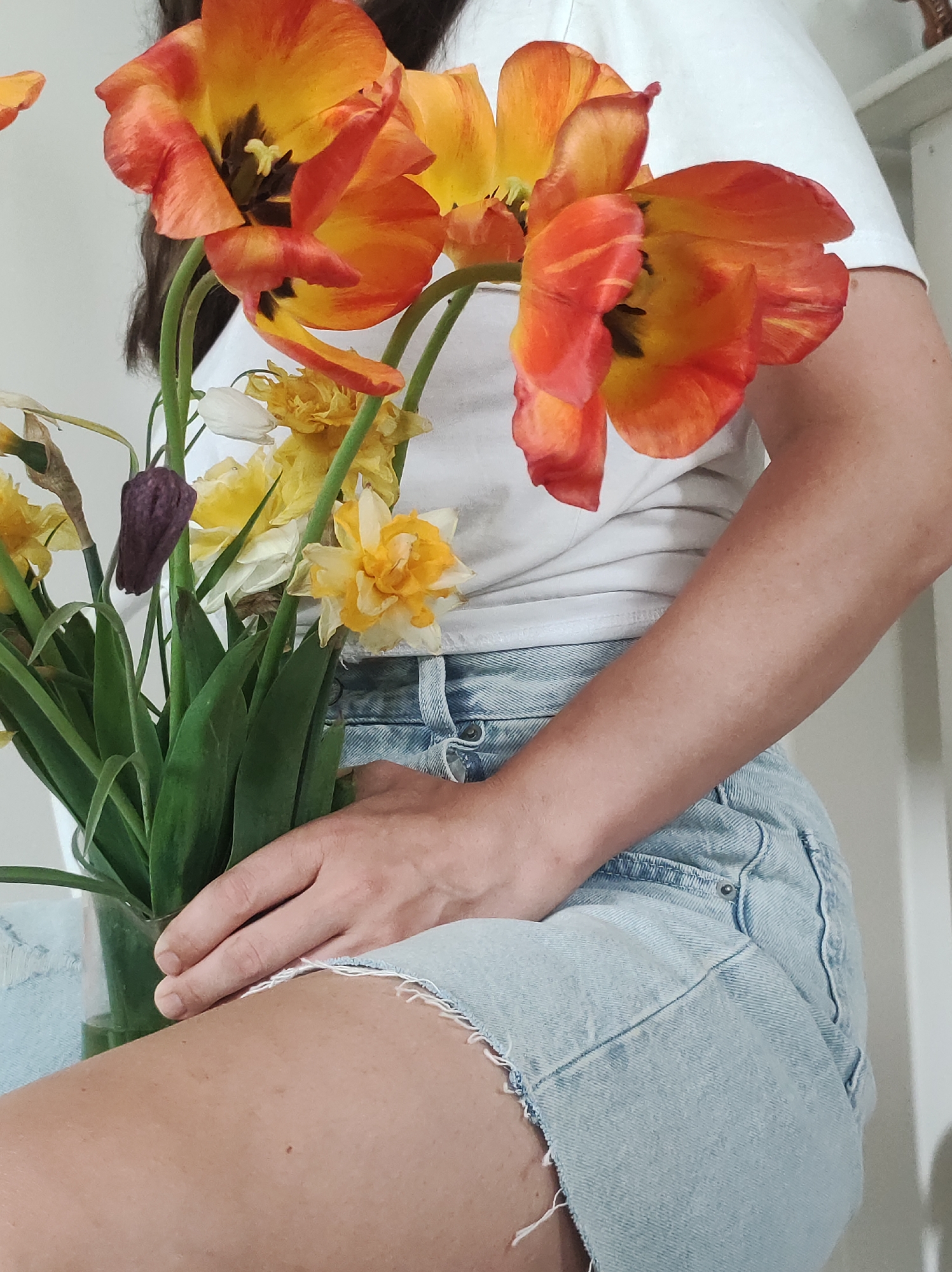 A person wearing a white t-shirt and light blue denim shorts is holding a glass vase filled with flowers. The flowers are a mix of orange tulips and yellow daffodils. The person's arm is visible on the right side of the image, and their hand is resting on the vase. The background is a plain white wall.