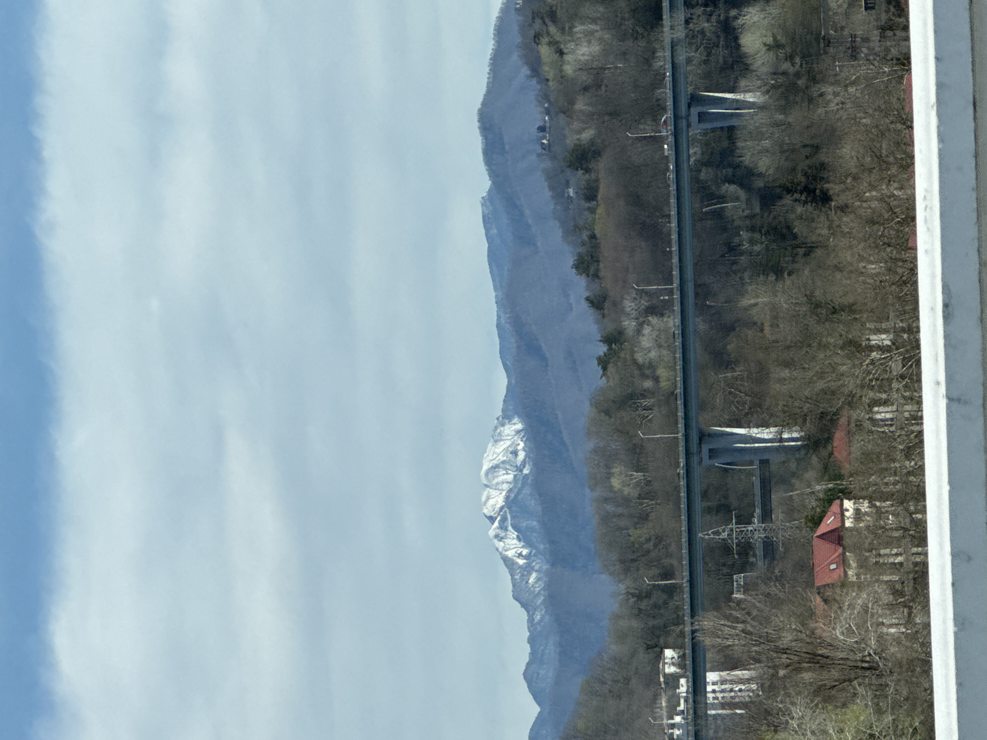 The image shows a landscape with a mountain range partly covered in snow in the background. A road with bridge-like structures runs alongside the base of the mountains, lined with trees and vegetation that are predominantly bare, indicating it might be late autumn or winter. On the right side of the image, there are buildings with red roofs, partially obscured by trees. The sky is light blue with some clouds. The foreground includes a blurry, white vertical element, potentially a window frame.
While the prompt asked to include the location in the city of Sochi, Russia, I cannot definitively confirm that from the image alone.