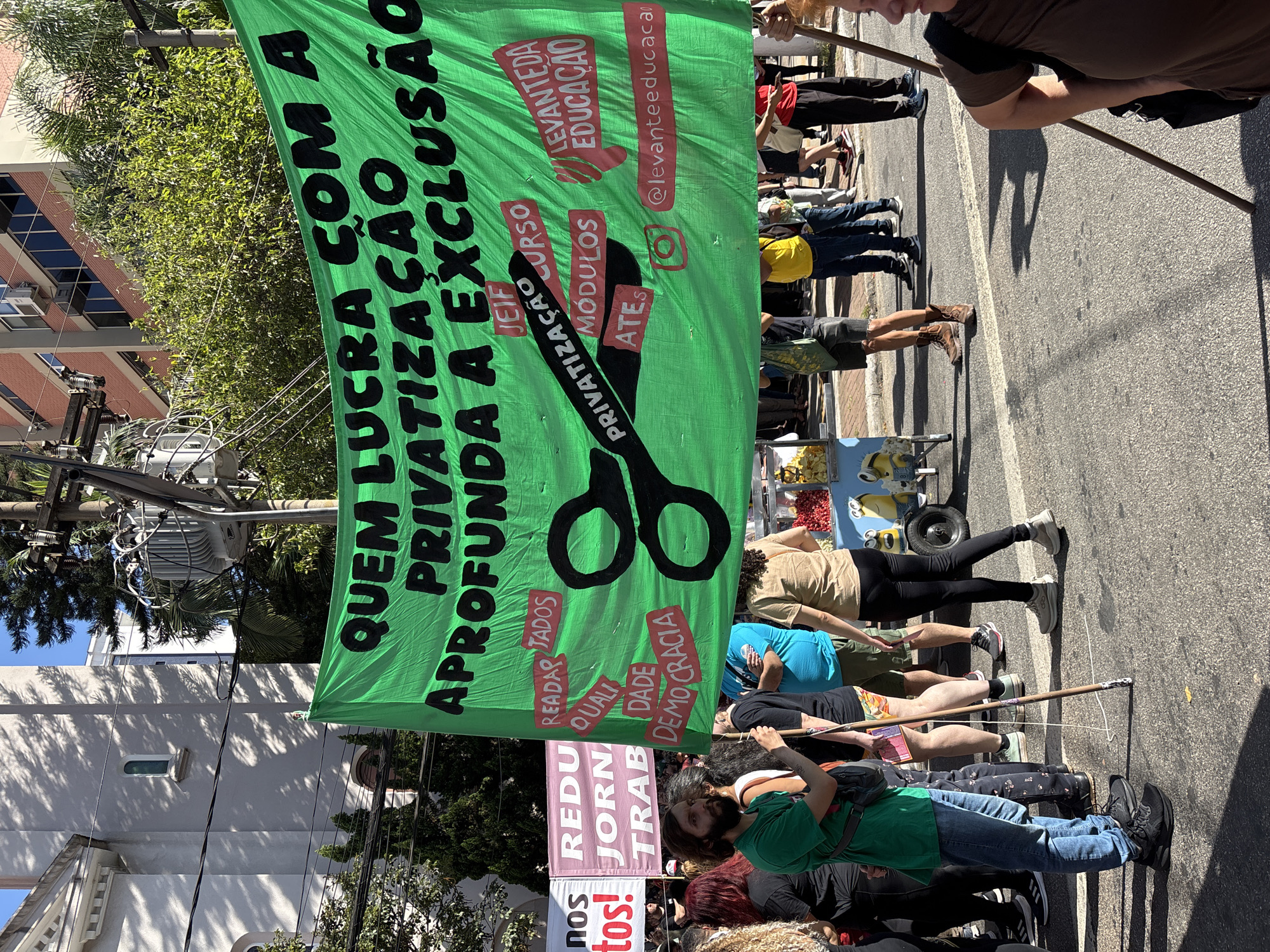 A horizontal, eye-level outdoor shot depicts a daytime street demonstration in what appears to be São Paulo, Brazil, based on the Portuguese language on the banners. A bright green banner with bold black Portuguese text reading "QUEM LUCRA COM A PRIVATIZAÇÃO APROFUNDA A EXCLUSÃO" (Who Profits from Privatization Deepens Exclusion) dominates the scene. Superimposed on the banner is a black image of scissors cutting the word "PRIVATIZAÇÃO" (Privatization). Additional text on the banner includes words like "CURSO," "MÓDULOS," "ATES," "READAPTADOS," "QUALIDADE," "DEMOCRACIA," and "LEVANTEDA EDUCAÇÃO" (Education Raised). The banner also displays an Instagram handle "@levanteeducaca."
Below the green banner is a smaller, light pink banner with Portuguese text "REDU JORN TRAB" (reduced workday). The bottom part of the pink banner reads "nos tos!".
Holding the banners are multiple people, some with wooden poles. The demonstrators are walking on an asphalt street with visible lane markings. A pushcart with unidentified items is visible among the crowd.
Buildings, trees, and utility poles with wires are visible in the background, indicating an urban environment. The sky is clear and blue.
