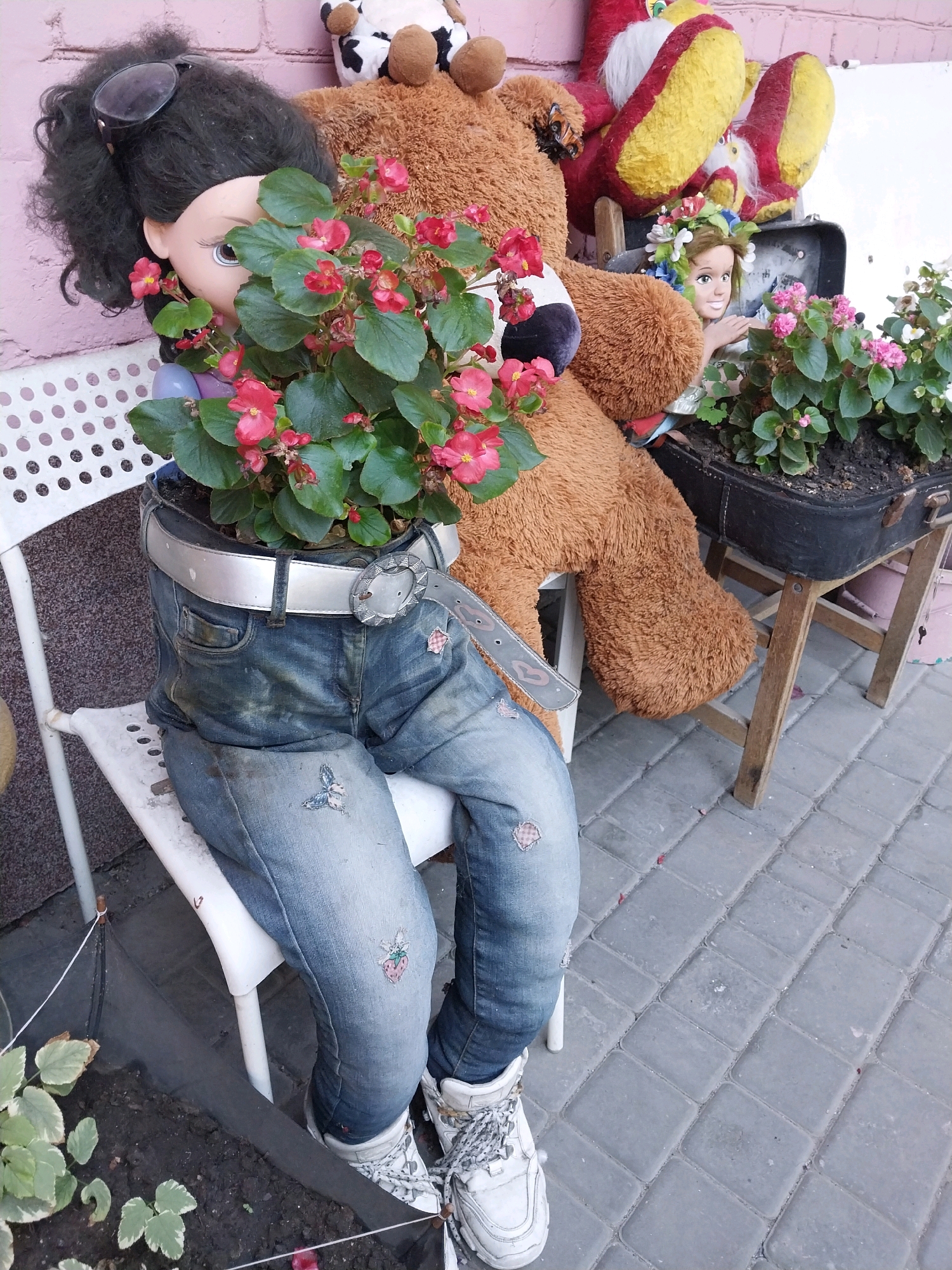 In Dnipro, Ukraine, a whimsical garden art installation is displayed outdoors. A large teddy bear, seemingly part of the display, is positioned behind a plant pot fashioned from a pair of denim jeans. The jeans are worn by a doll's torso, which is protruding from the top of the pot, partially obscured by red-flowered plants. The doll wears dark, curly hair and sunglasses pushed up onto her head. Her exposed face shows a light skin tone and large brown eyes. The jeans are belted with a silver belt. The overall scene suggests a creative and unconventional use of materials for garden decoration.