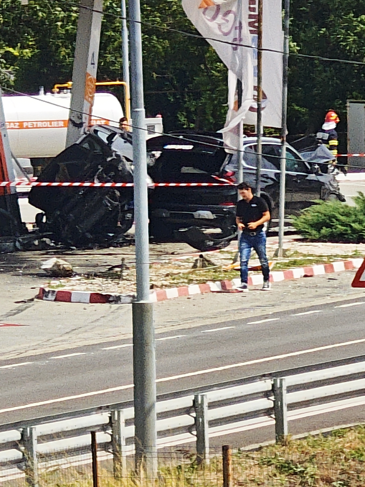 The image captures the aftermath of a severe traffic accident near Coșoveni, Romania. In the foreground, a man in a black polo shirt and ripped blue jeans walks away from the wreckage, looking down at his phone. The scene is cordoned off with red and white striped tape. Behind the tape, a heavily damaged black car lies on its side, its front end crumpled and distorted. A large white tanker truck, labeled "GAZ PETROLIER", is partially visible behind the car. In the background, a person in a yellow and black uniform, likely a firefighter, stands near the truck. The environment appears to be an intersection or roadside, with trees and foliage visible in the distance. The overall impression is one of chaos and damage.