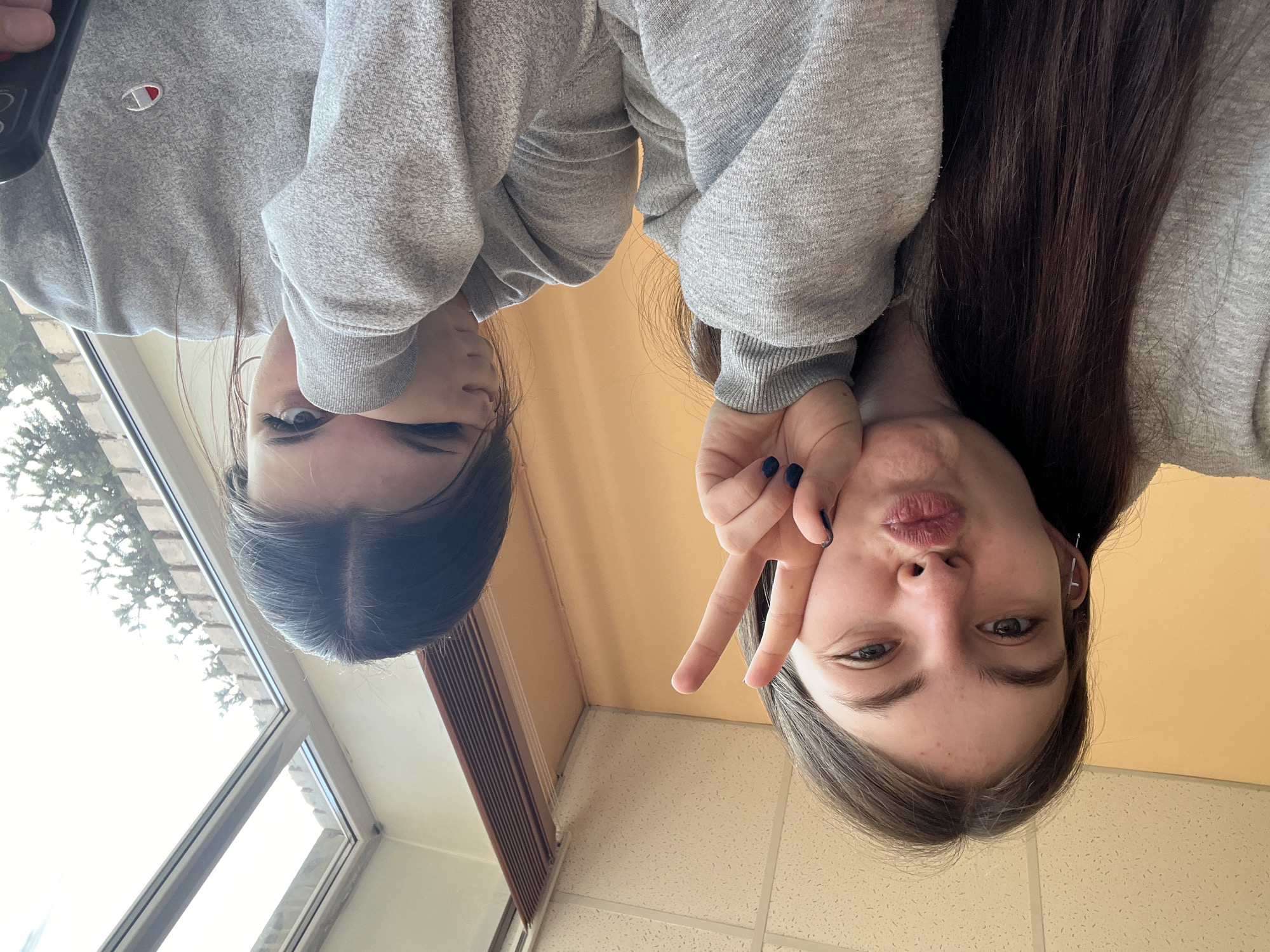 Two young women wearing gray sweatshirts are posing for a selfie indoors. The woman on the left has dark brown hair, and the woman on the right has lighter brown hair. The woman on the right is making a peace sign with her right hand and puckering her lips. The environment appears to be an indoor space with a window and a wall. The location in the city of Saint Petersburg, Russia cannot be determined from the image.