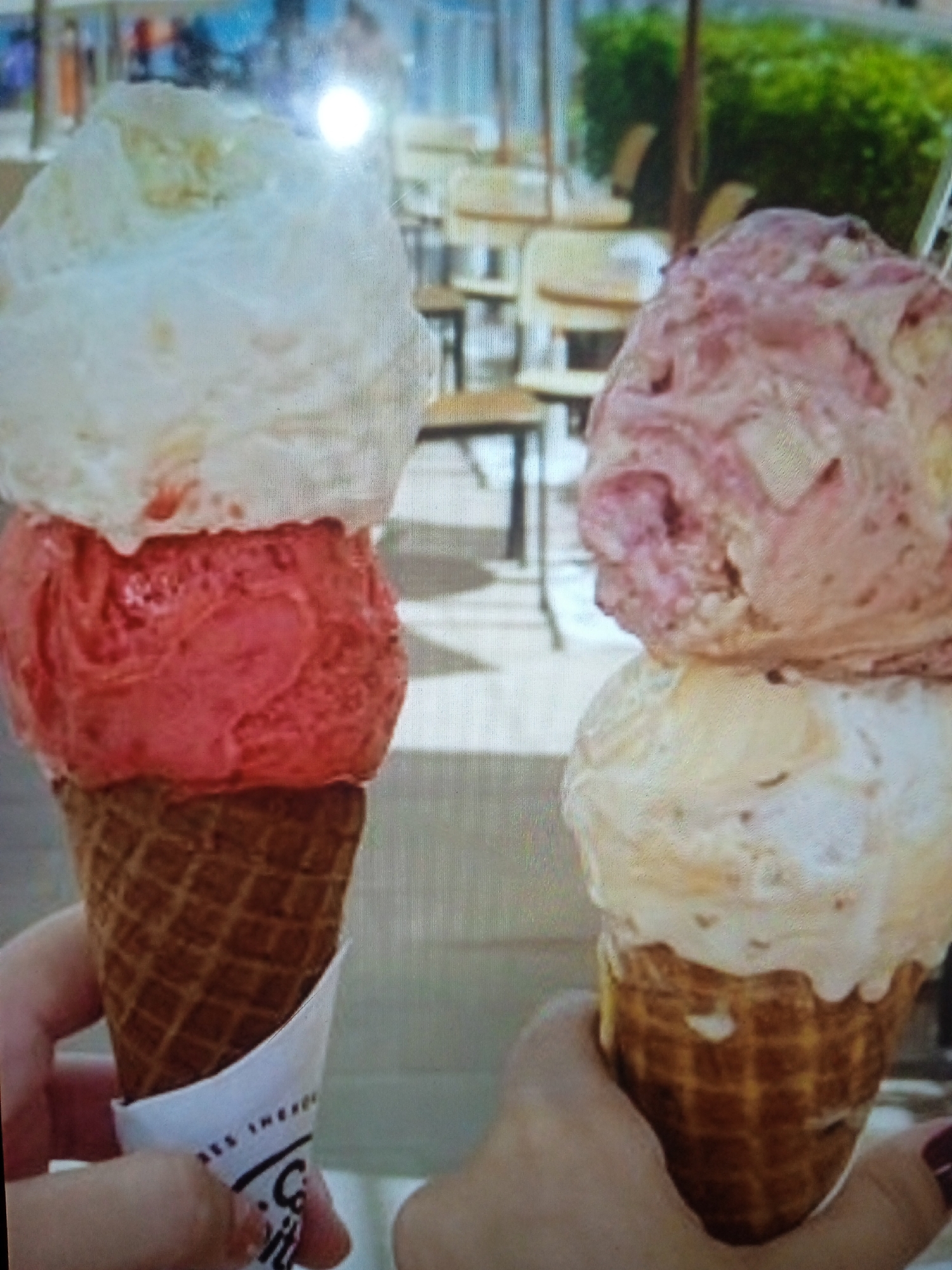 The image shows two ice cream cones. The cone on the left has three scoops of ice cream: white on top, red in the middle, and a portion of the waffle cone is visible at the bottom. The cone on the right has two scoops: a pinkish-red on top and a lighter color, possibly vanilla, below. Both cones appear to be in a seated outdoor dining area with chairs and tables in the background, possibly a cafe or restaurant. The image is somewhat blurry and overexposed in places, obscuring finer details of the surroundings and the ice cream flavors. There are no visible people clearly identifiable, though hands are holding the cones. There is no specific information in the image that would allow for identification of the location within the city of Chernivtsi, Ukraine.