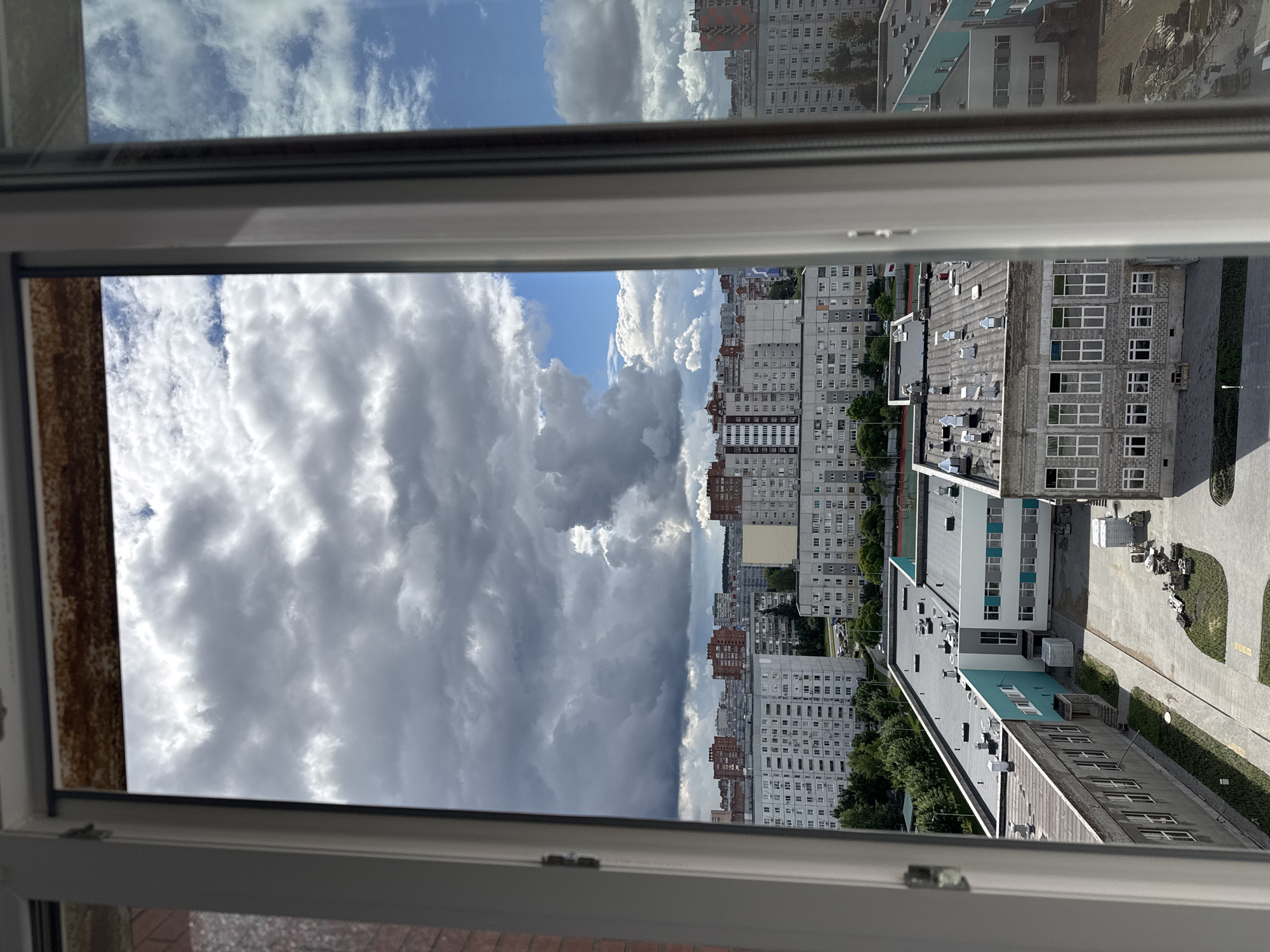 The image, captured from a window and presented with a 90-degree clockwise rotation, displays an expansive urban landscape beneath a dramatic sky.
In the foreground, a clean white window frame encloses the view, with a section of rusted, textured brown material visible along the left edge, possibly part of an older frame or sill.
Beyond the window, the scene unfolds into a dense city environment characterized by numerous multi-story buildings. A significant portion consists of large, light-colored residential apartment blocks, some with repeating window patterns and occasional red or blue-tinted facades. Lower-rise commercial or industrial buildings with flat roofs, many equipped with ventilation systems and air conditioning units, are also visible. Ground level areas show paved roads, sidewalks, and patches of green space with trees and grass separating the structures.
The sky is a prominent feature, displaying a striking contrast of cloud formations. Heavy, dark grey clouds mass together, suggesting overcast or stormy weather, while bright white cumulus clouds are interspersed throughout. Patches of clear blue sky are visible, particularly towards the upper right, providing glimpses of sun. The lighting suggests it is daytime, likely mid-morning or afternoon, with moderate shadows cast by the buildings. No individuals or specific activities are discernible within the urban environment, and no visible text is present.